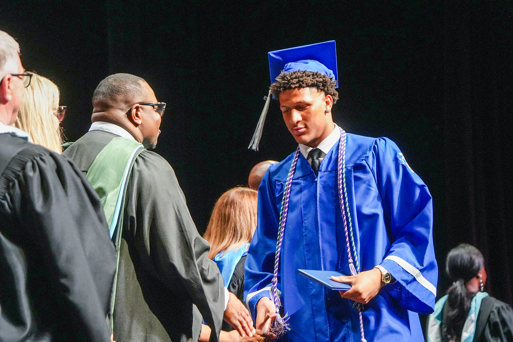 A young person in a blue graduation gown and cap shakes hands with an adult in a black gown.