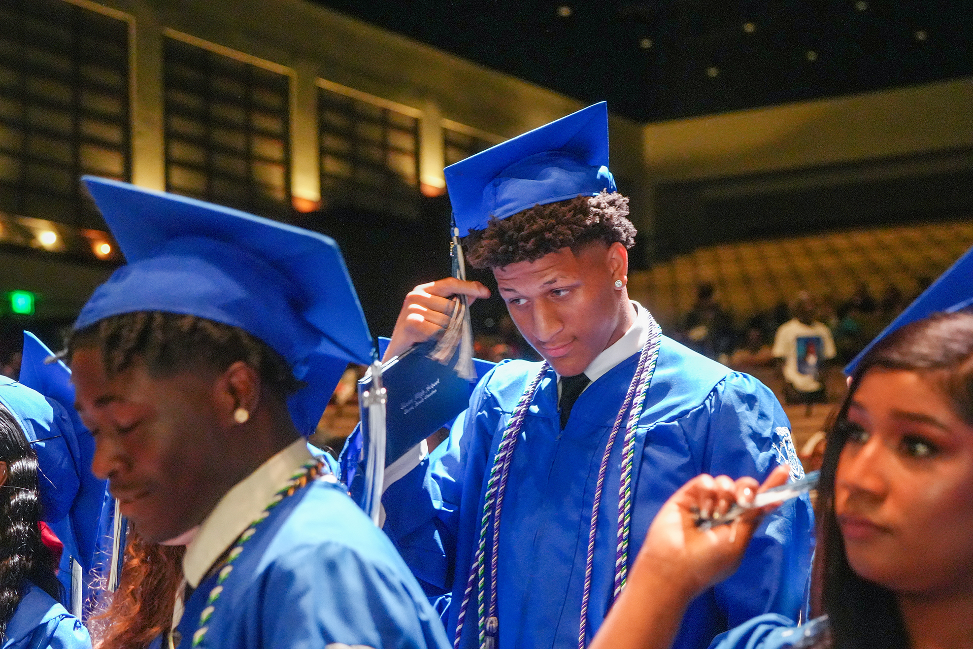 A group of students in blue gowns and caps, two males and one female, in a large room.