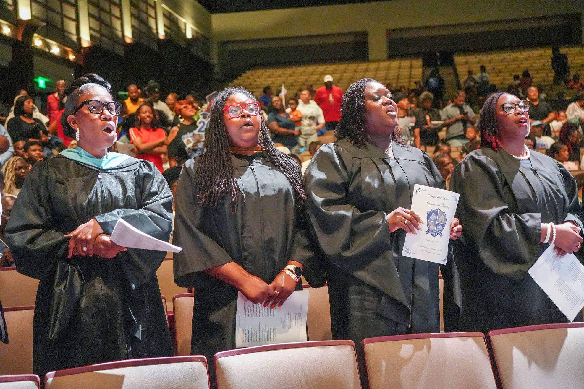 Four women in black gowns and glasses stand side by side holding papers, facing a large audience in an auditorium.