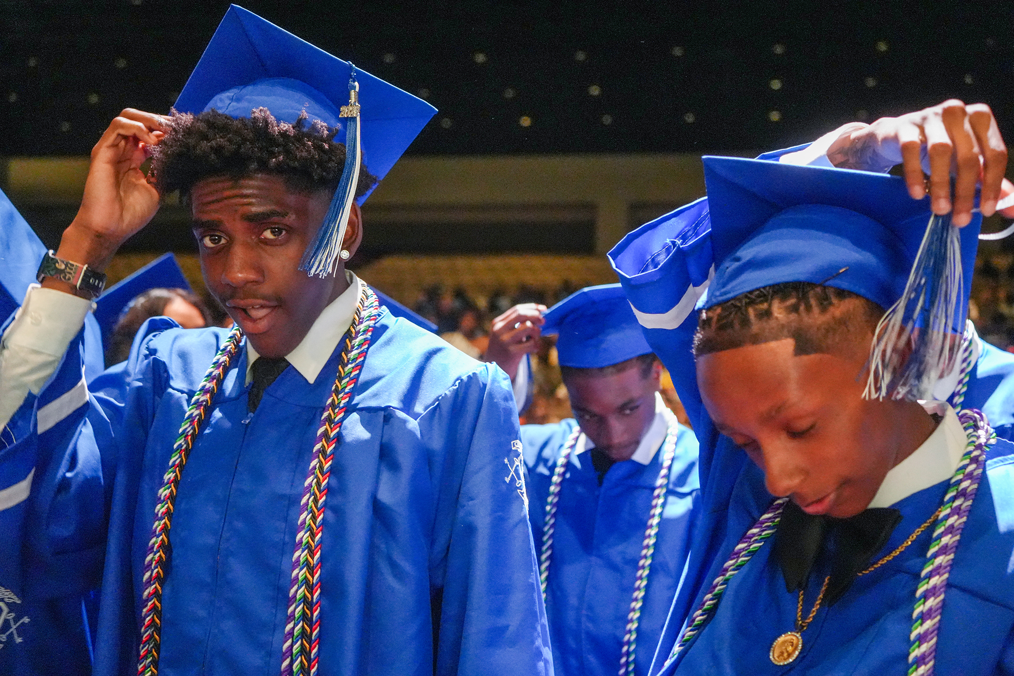 Several people in blue caps and gowns with tassels, adjusting caps. Background is blurred.