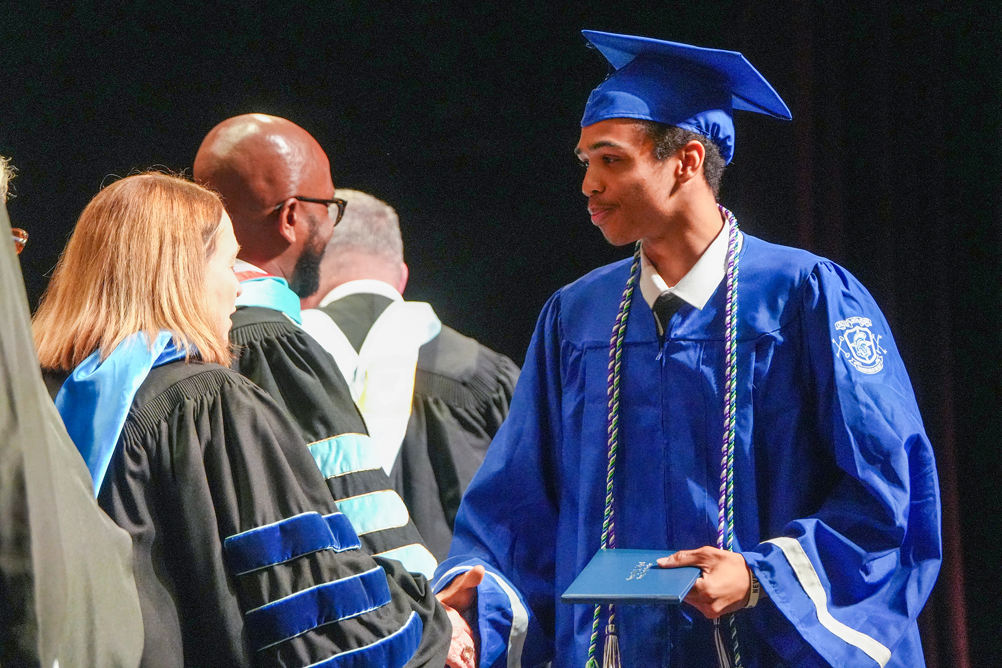 A man in a blue gown and cap greets someone during a ceremony, with three other adults in academic attire nearby.