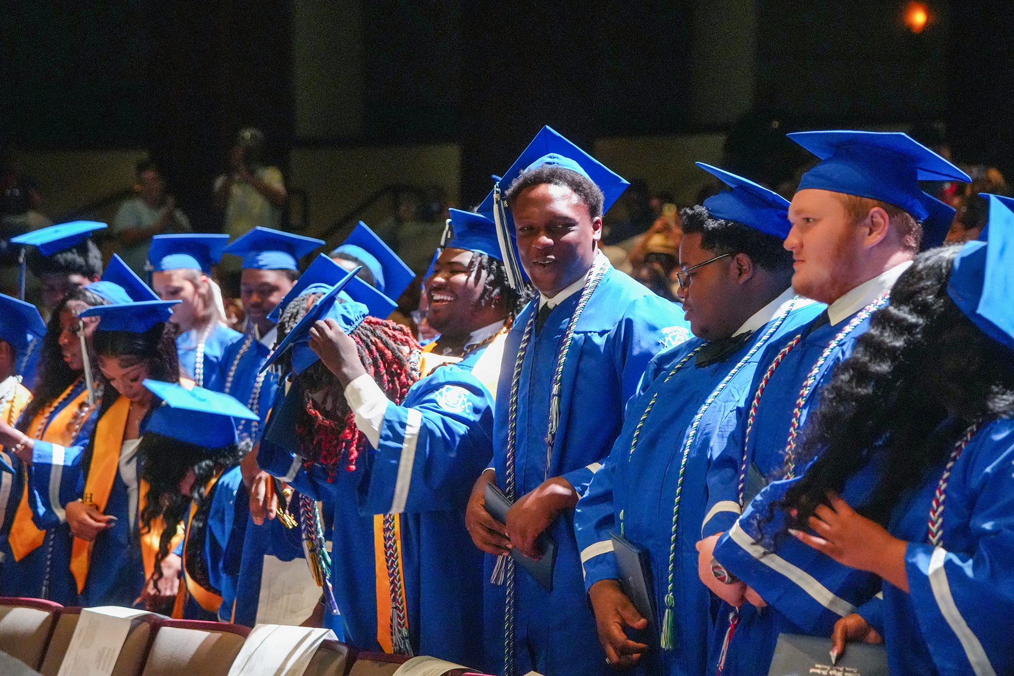 Graduates in blue gowns and caps stand in a row, some looking at the audience, in an indoor auditorium setting.