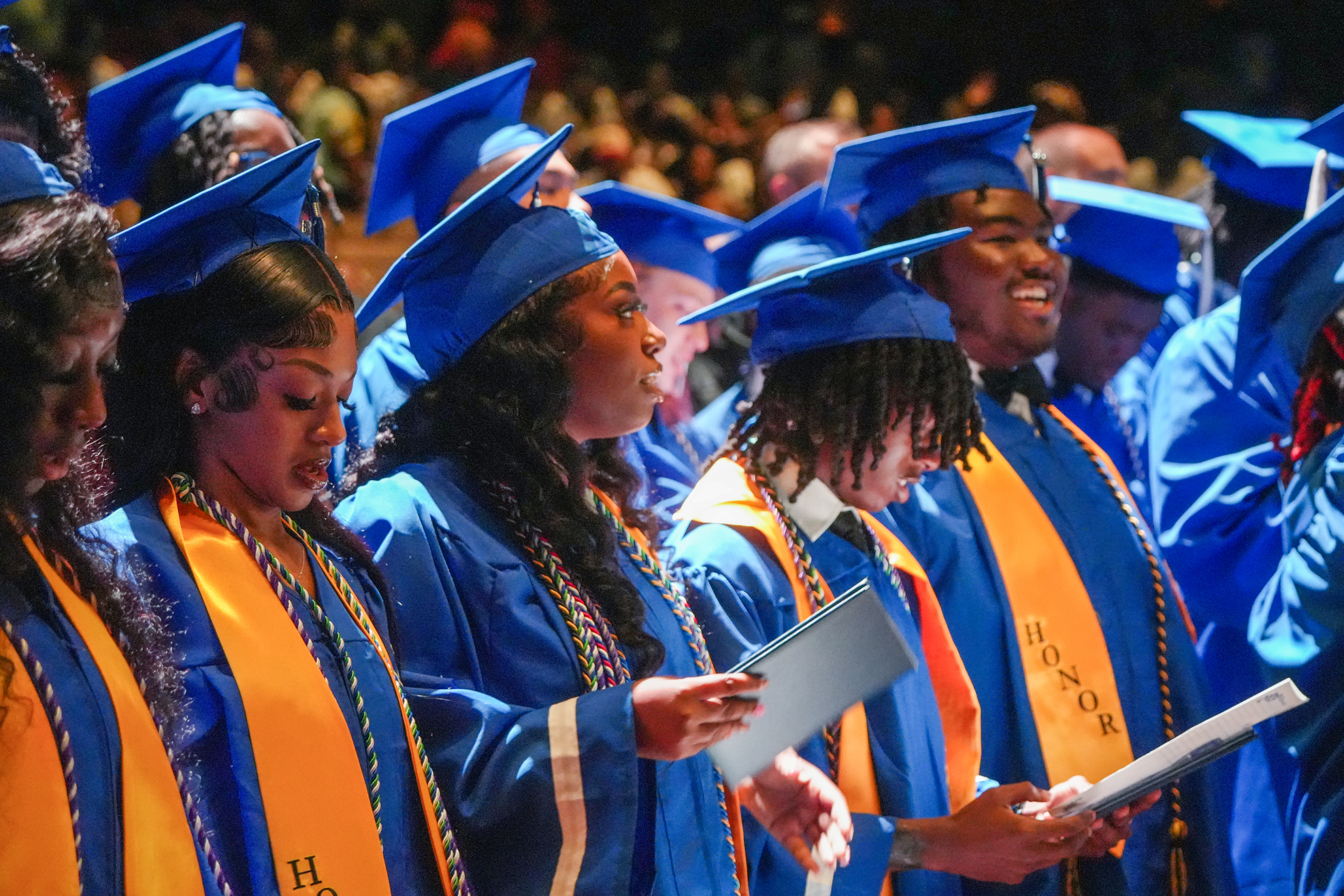 Graduates in blue gowns and hats sing in a ceremony. They hold books and wear orange sashes.