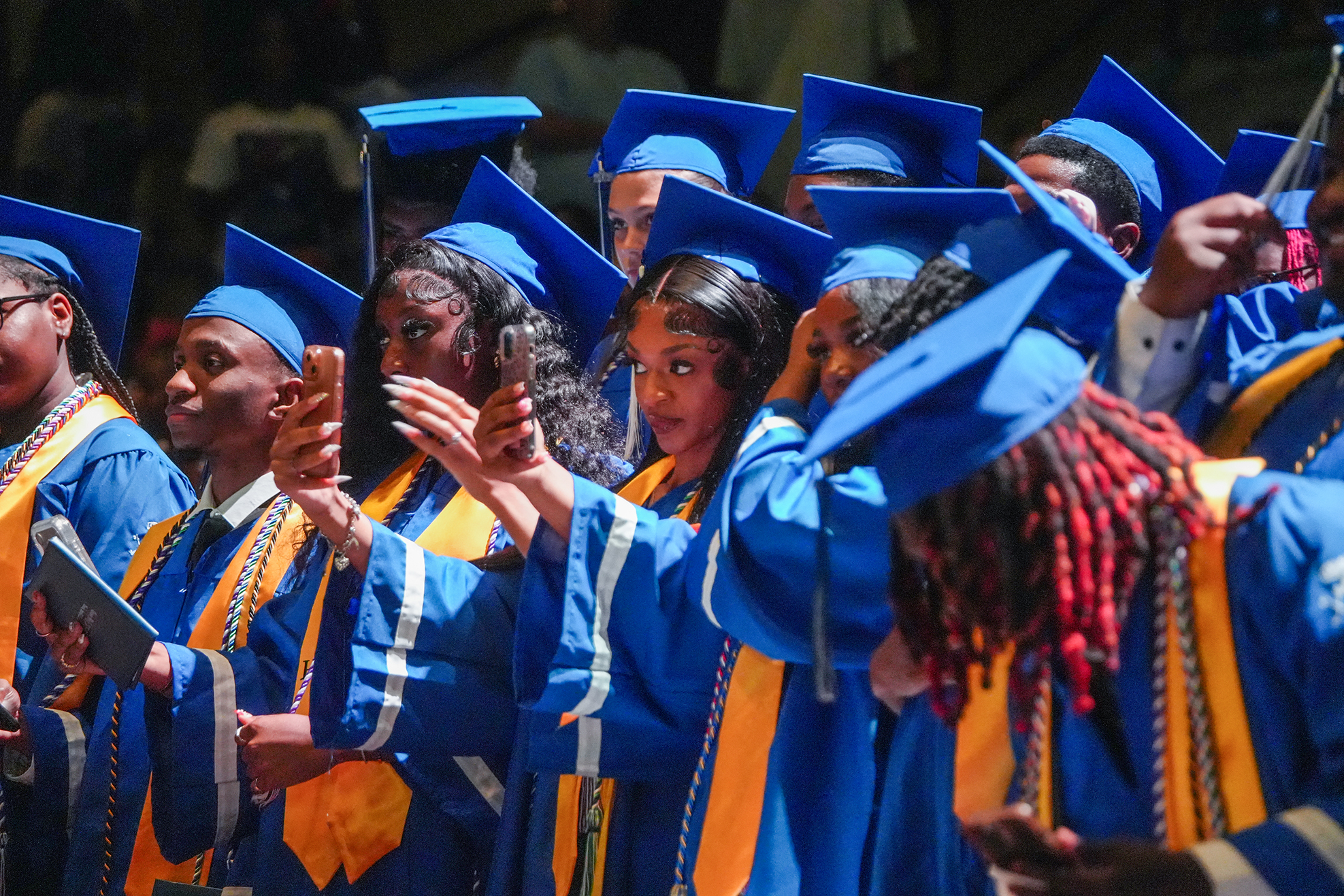 Graduates wearing blue gowns and caps stand in line, some with phones capturing the moment.
