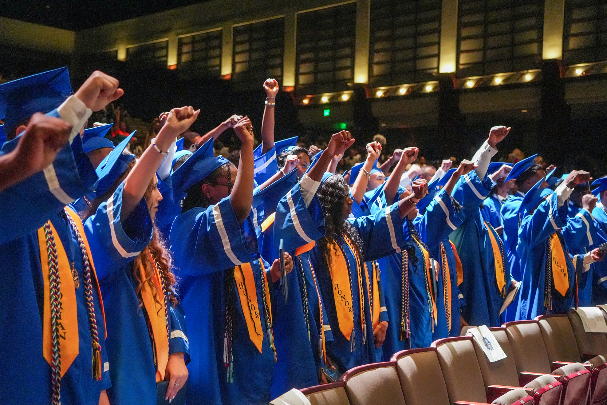 A group of people in blue gowns and caps stand in a row, raising their arms. Audience chairs are in the background.