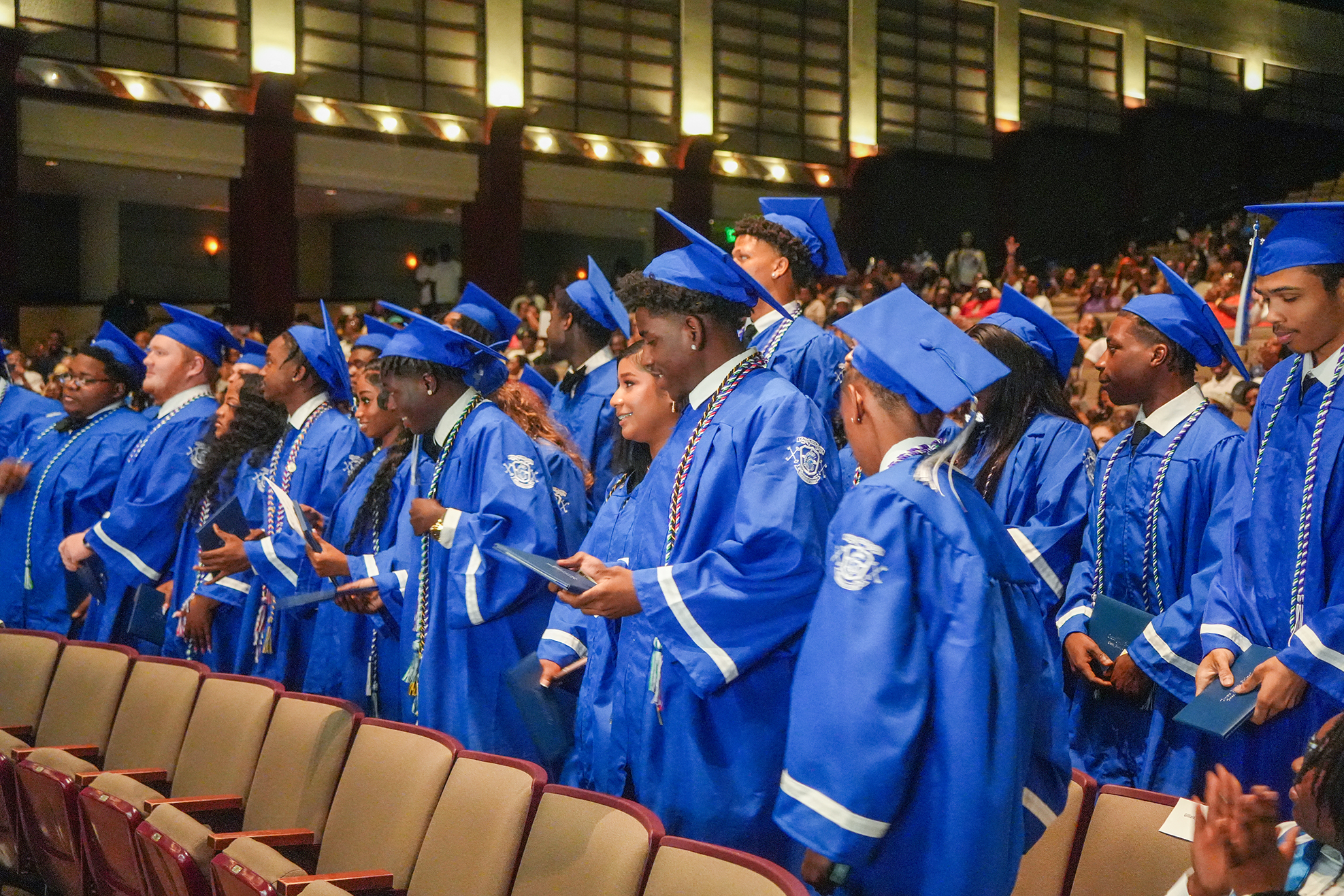 Graduates in blue gowns and caps stand in rows, facing forward, at a ceremony in an auditorium.