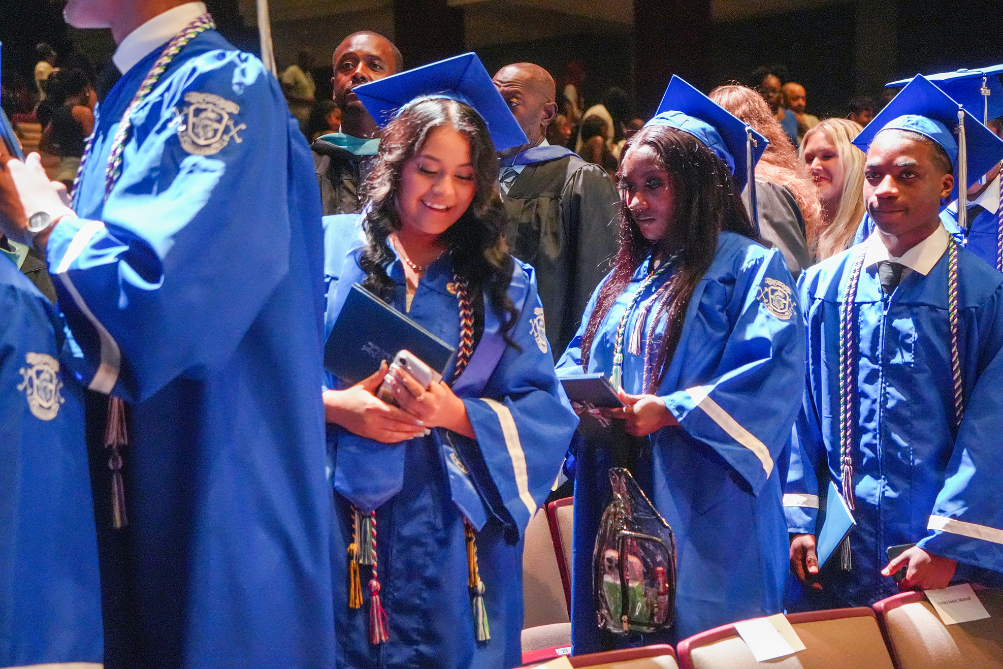A group of people in blue graduation robes, holding books, standing in a row and looking at something.