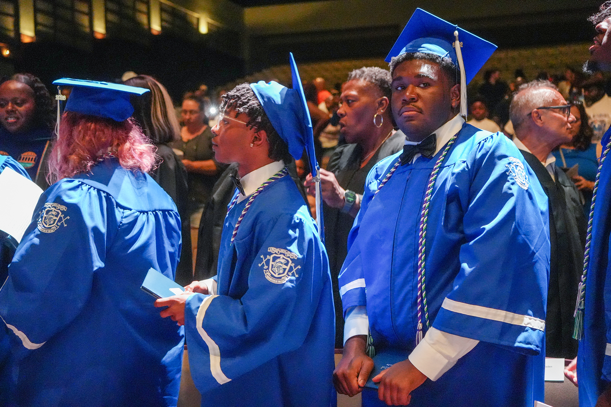Graduates in blue gowns and caps stand in a row. Behind them, a crowd watches from an audience.