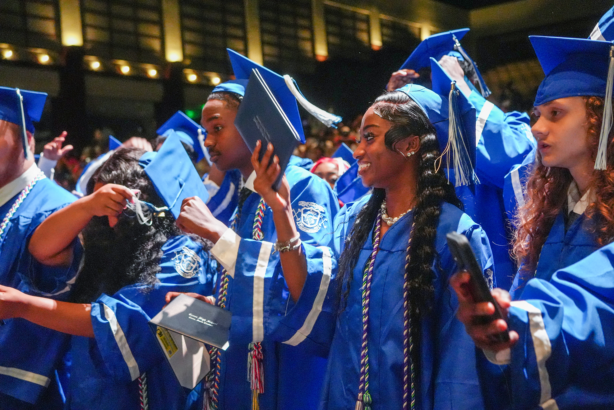 A group of graduates in blue gowns and caps stands in a room with blurred audience members.