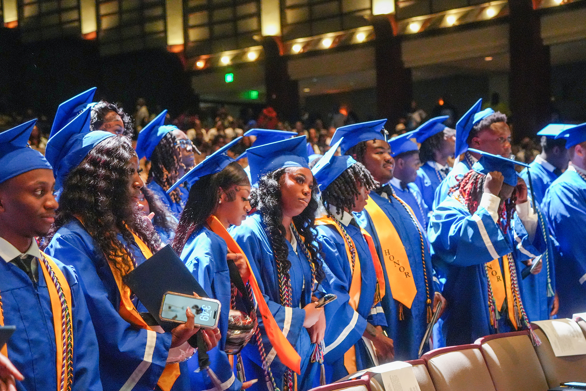 Graduates in blue and gold gowns and caps stand in rows, facing forward in an auditorium.