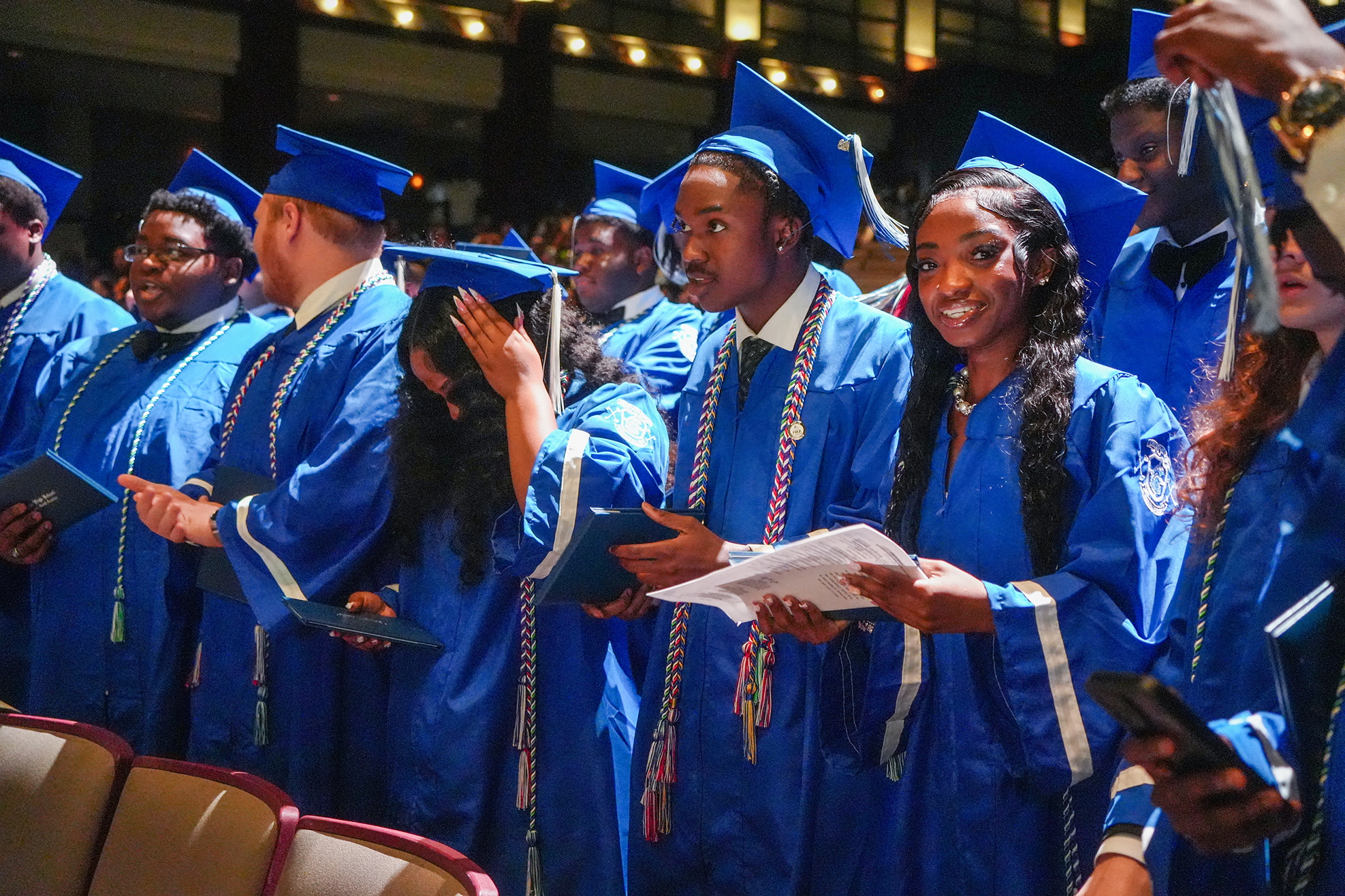 Graduates in blue gowns and caps stand in a line, holding diplomas, some reading papers, in a hall.