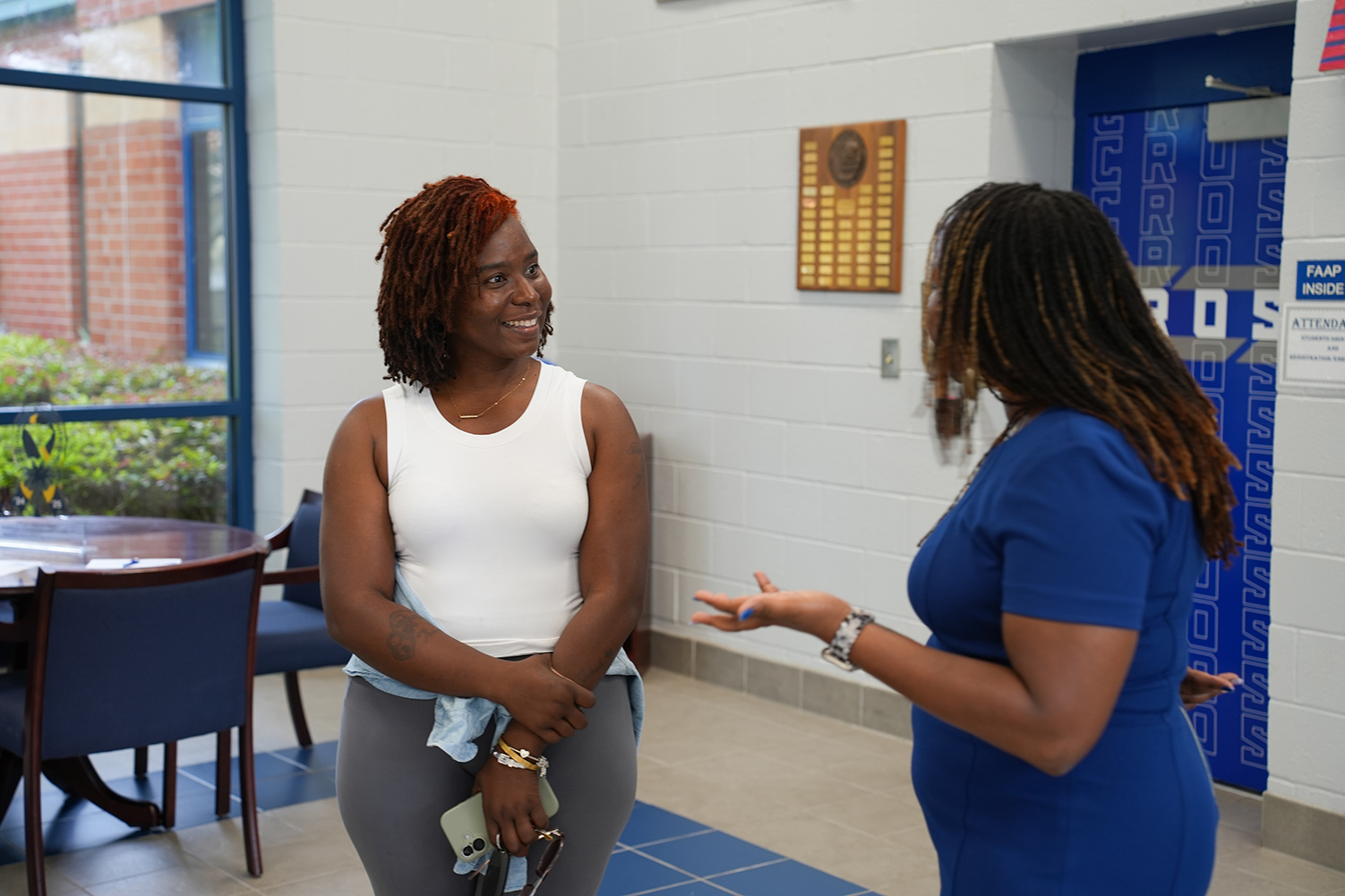 Two women converse in a hallway with a blue wall and table, while a blue door is visible in the background.