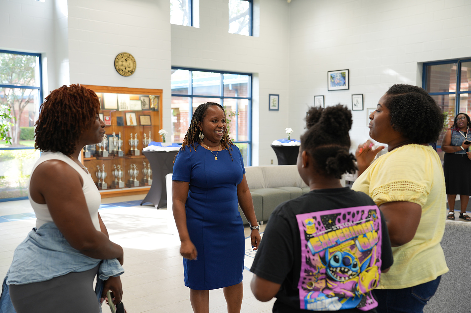 Four people standing and talking in a room with glass windows. One woman wears a blue dress, another a yellow shirt, and two more in casual wear.