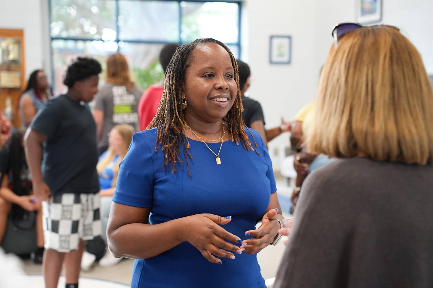 A woman in a blue shirt speaks to a woman in a gray sweater in a room with other people.