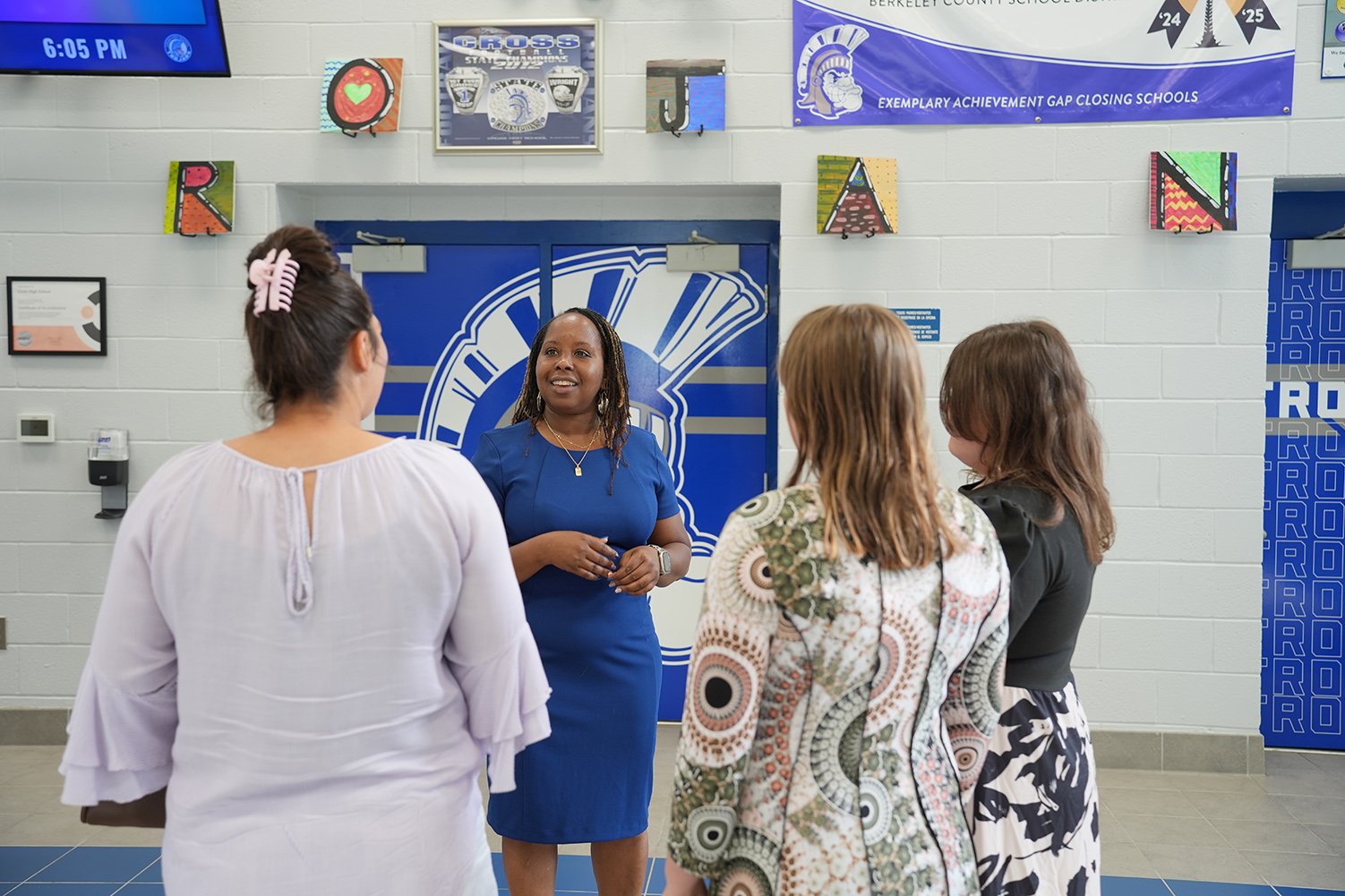 Four women stand in a room with blue doors. One woman in blue stands between two others in white and black.