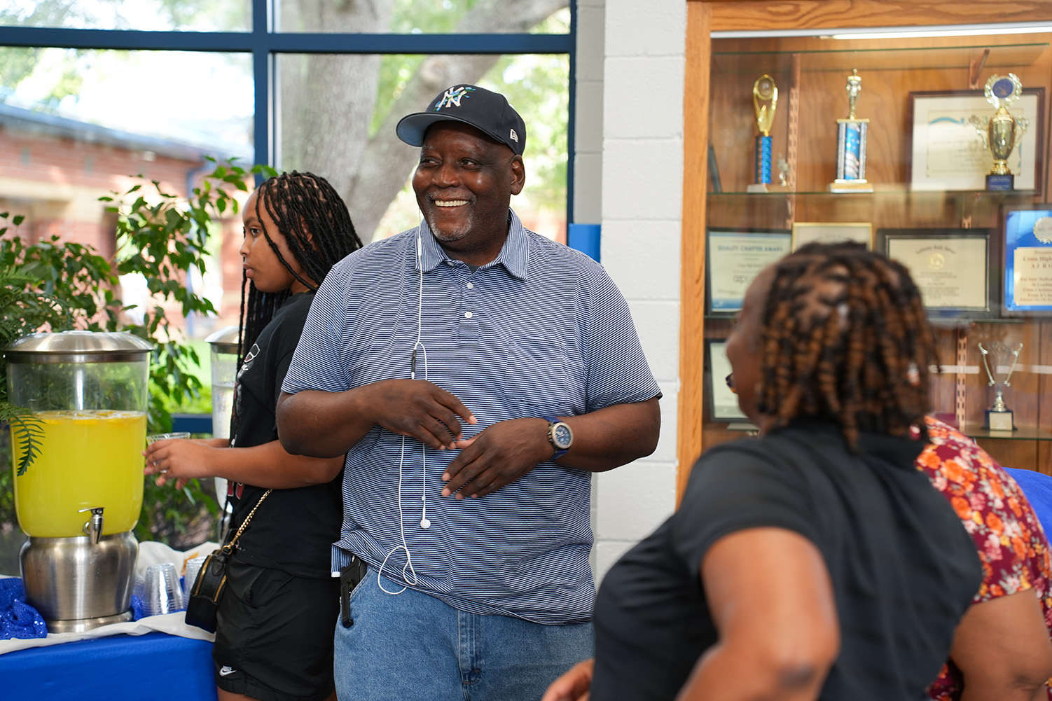 A smiling man in a blue shirt converses with two women. A juice blender is on a table.