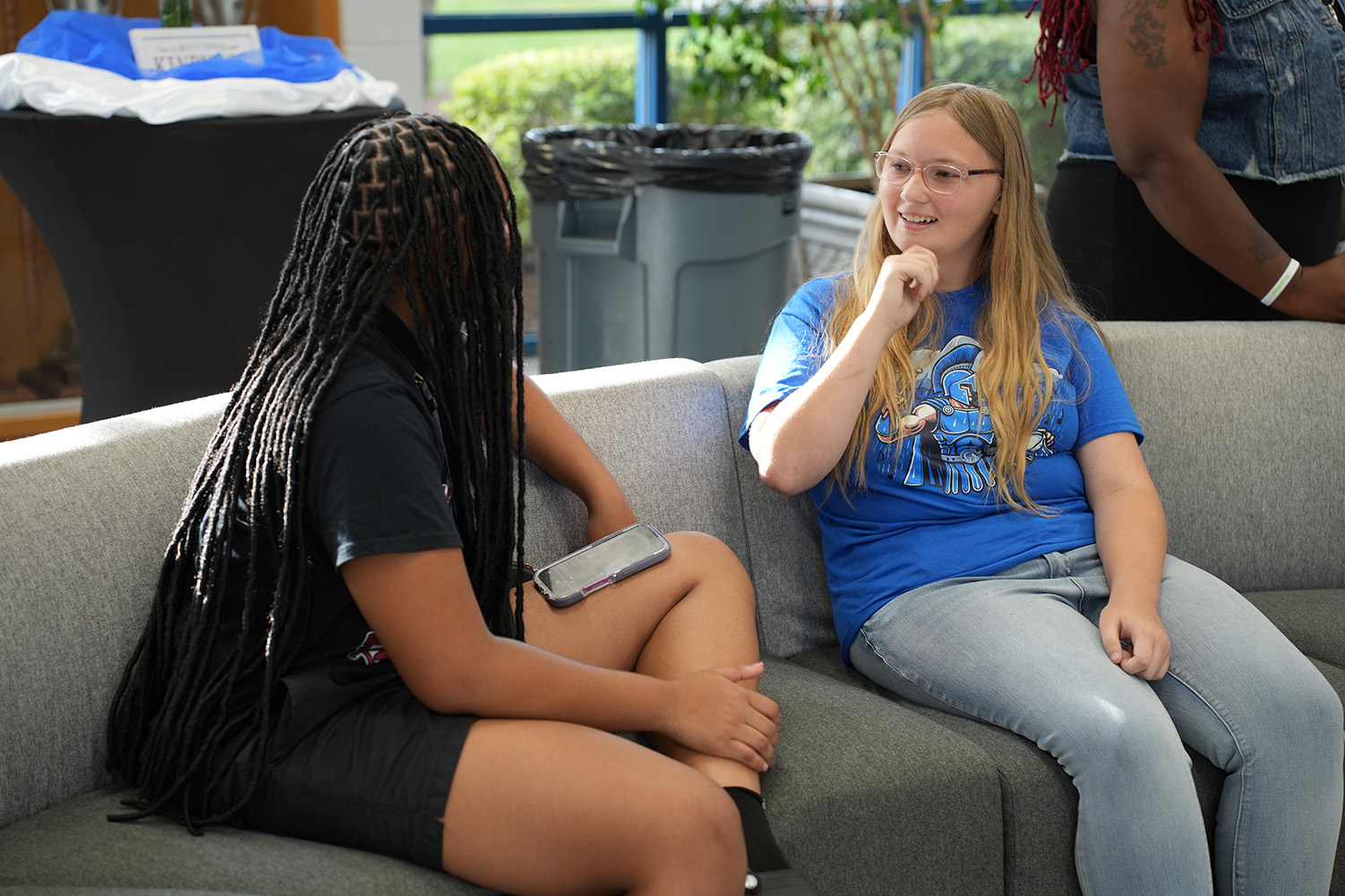 Two women sit on a couch in a room with a window, trash bins, and a plant. One wears glasses and a blue shirt, the other a black shirt.