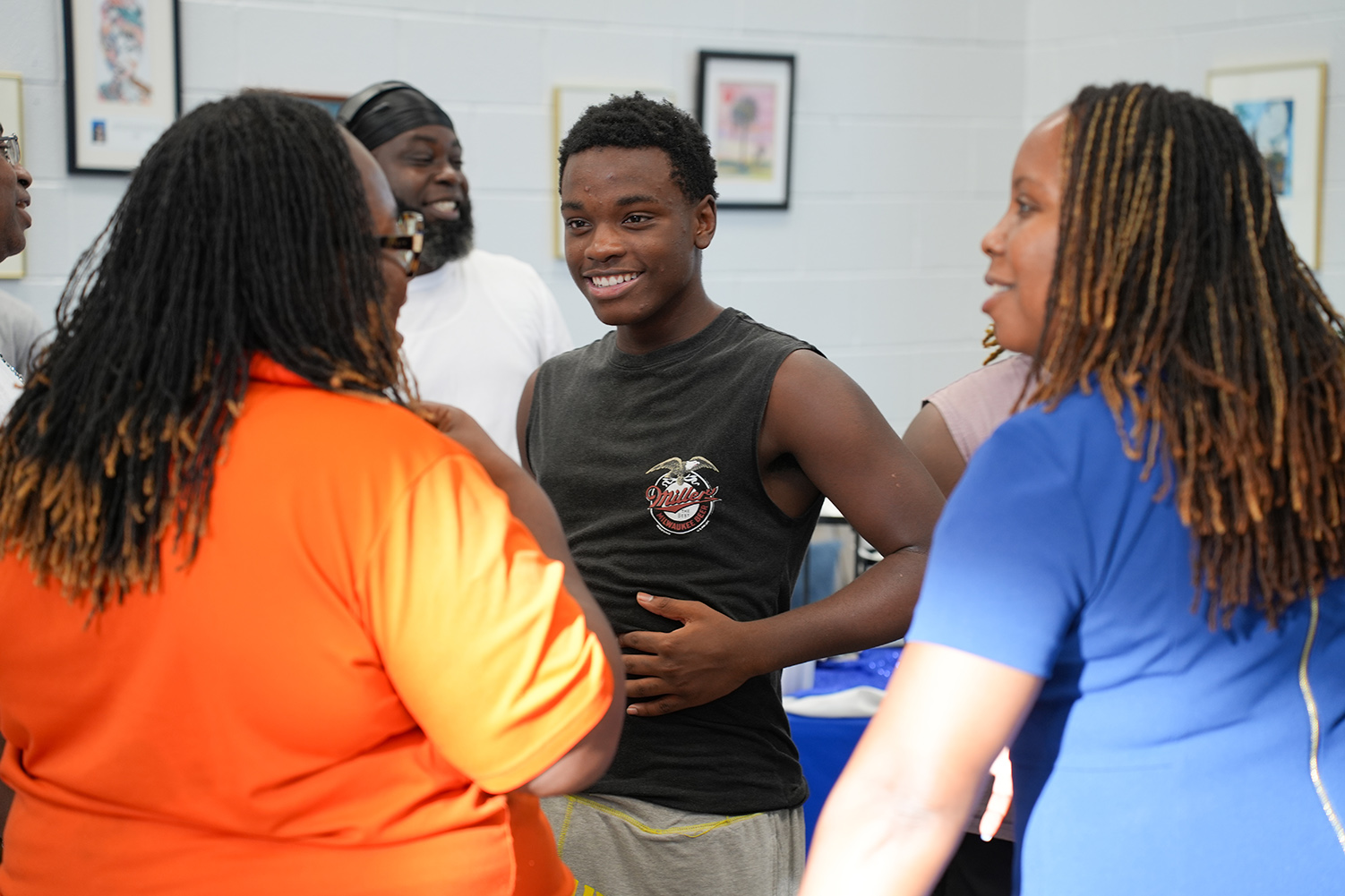 A group of people stands in a room. Two women and a man engage in conversation. The man wears a black sleeveless shirt.