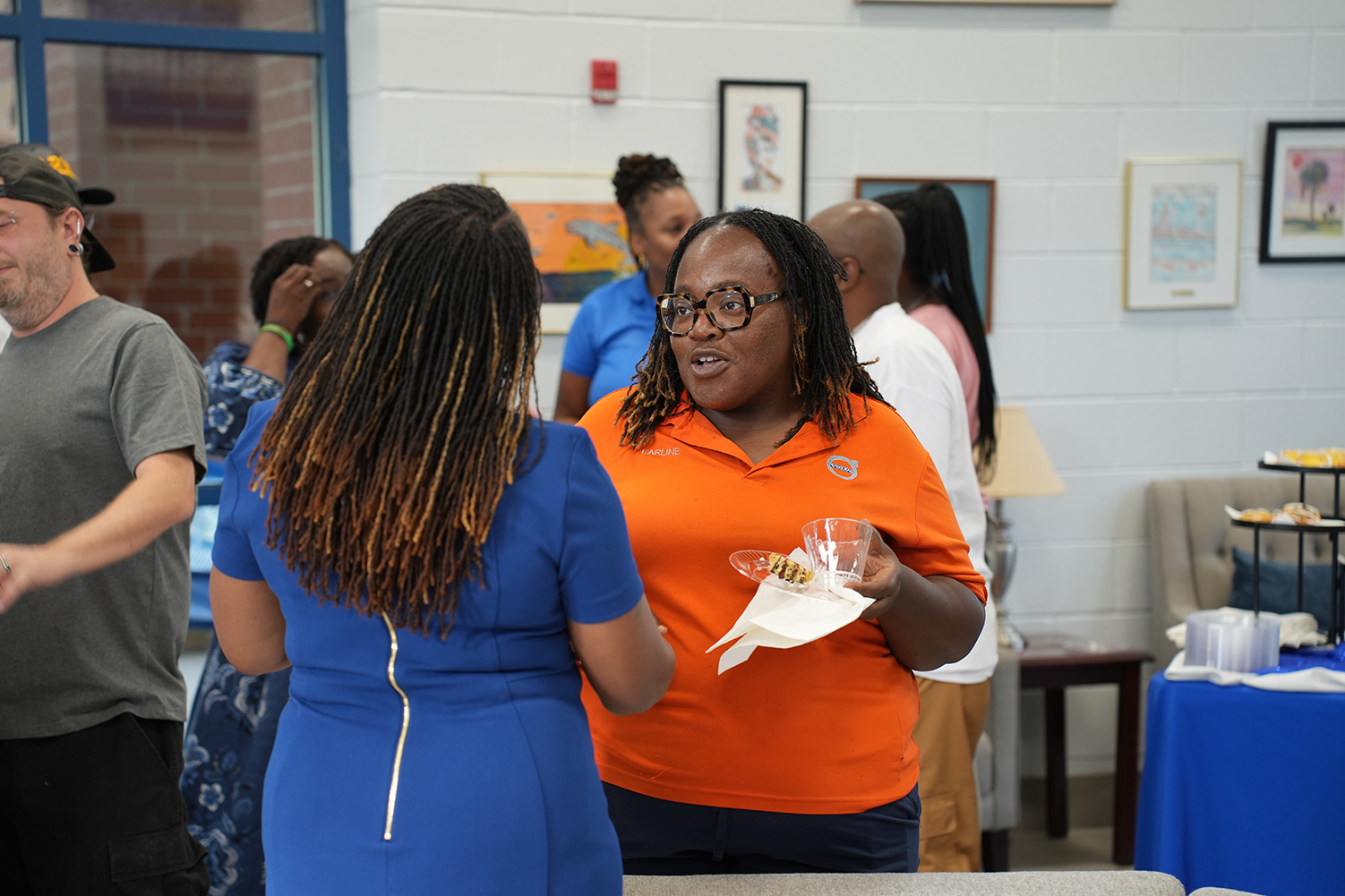 Several people in a room, two women converse with one holding a paper cup and napkin.