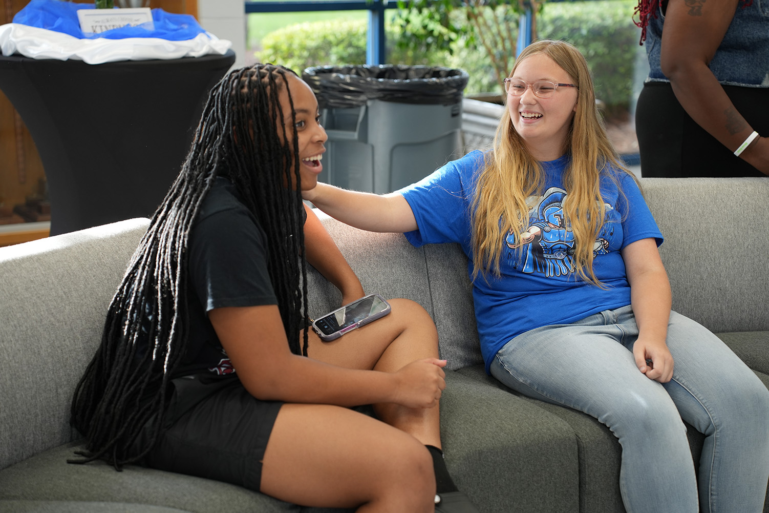 Two women sit on a couch; one with dark braids and one with blonde hair. One holds a cell phone.