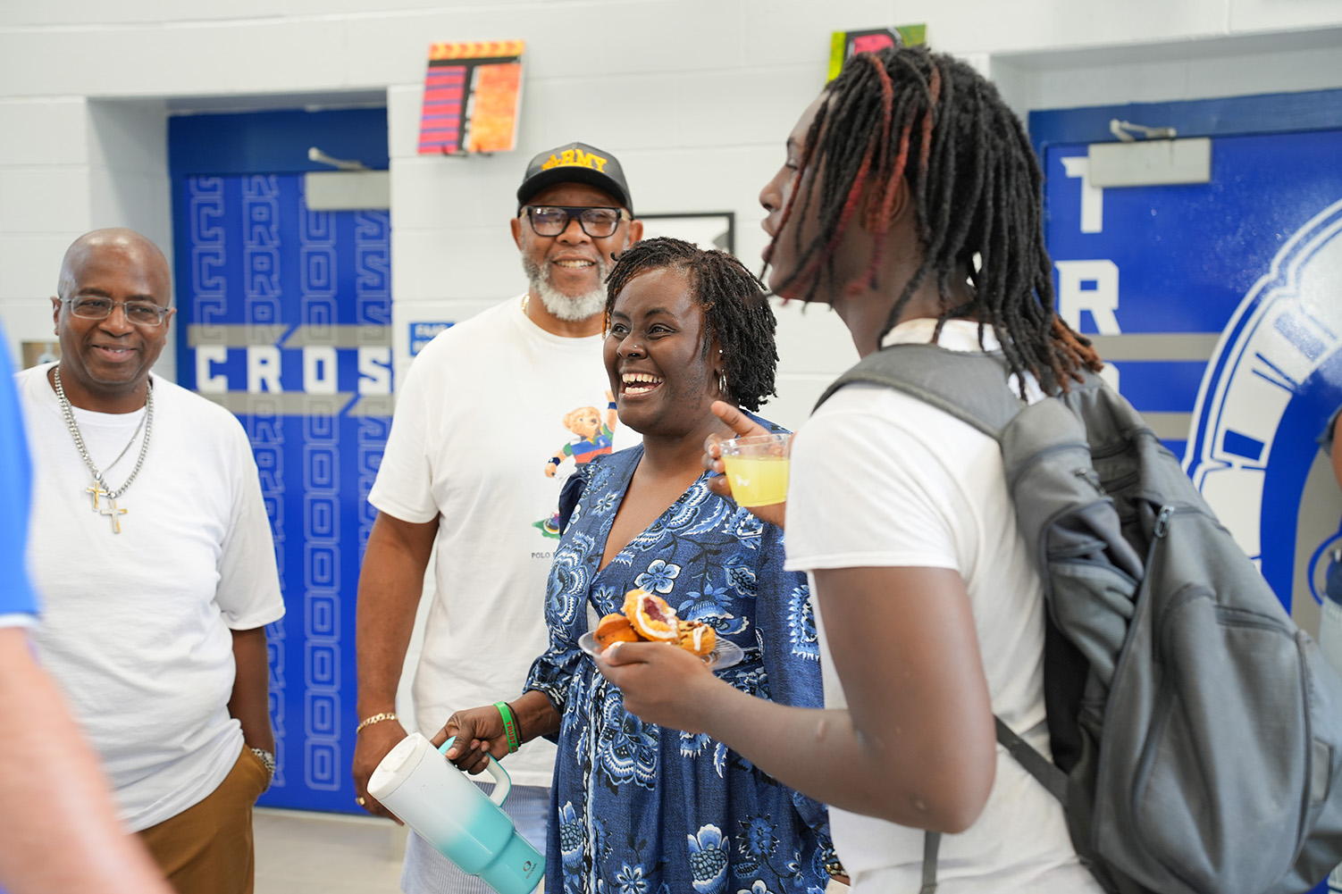 Four adults stand in a hallway with blue doors. Two of them hold drinks. One wears a backpack.