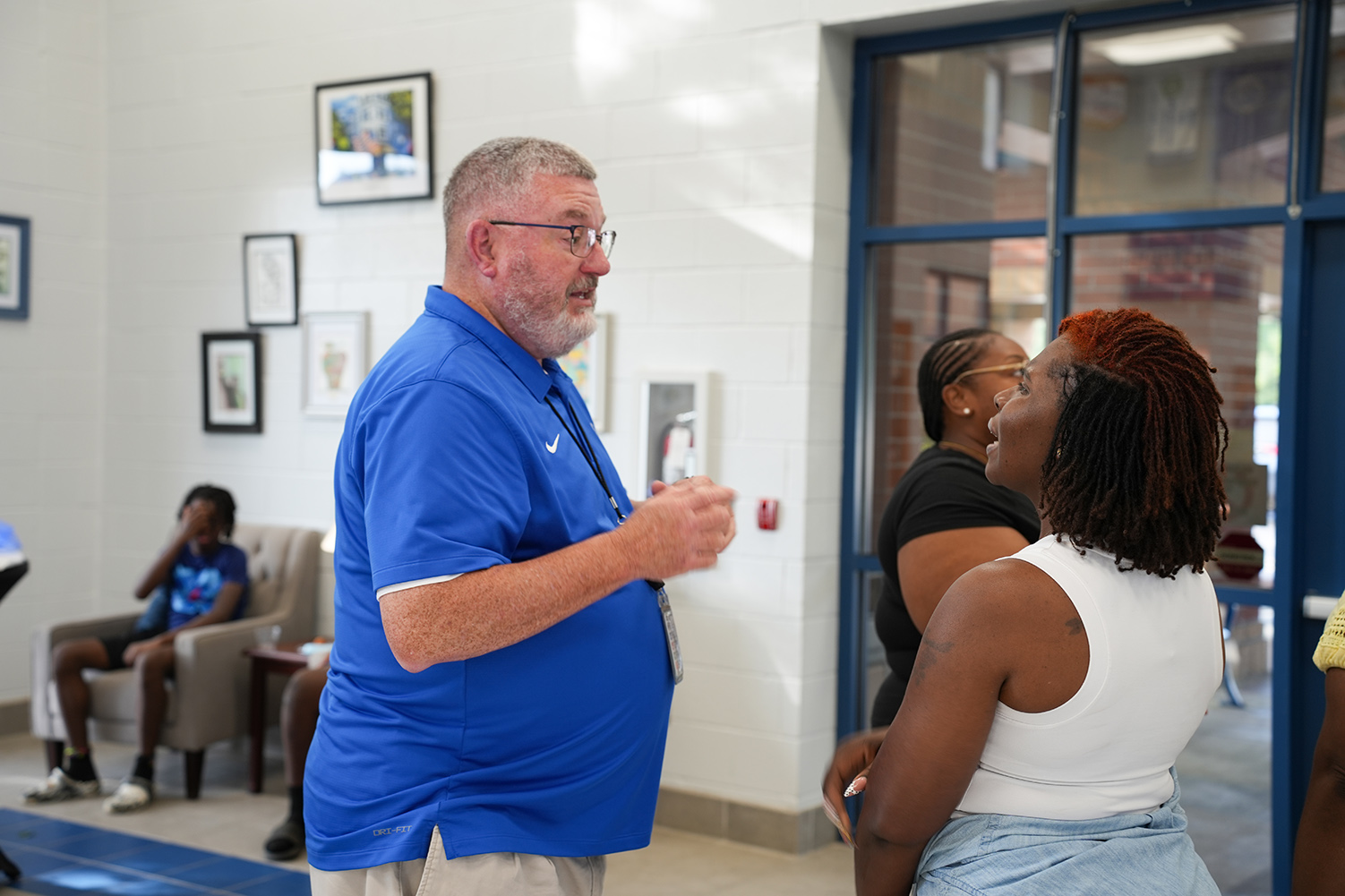 A man in a blue shirt is speaking with a woman with curly hair in a room with chairs and picture frames.