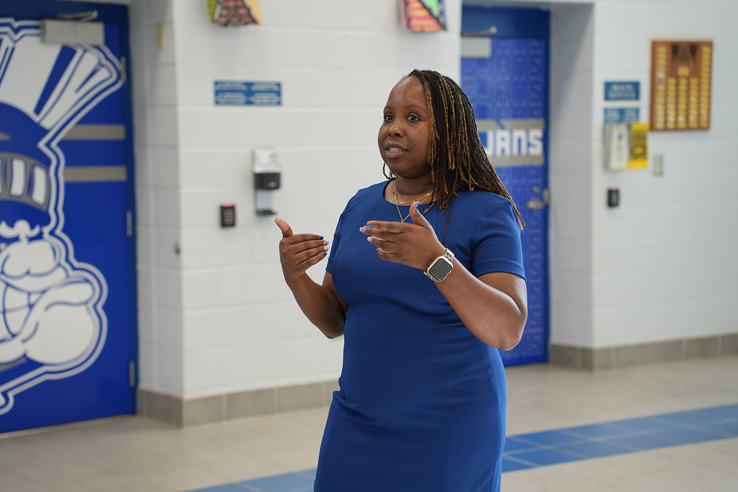 Woman with curly hair wearing a blue dress and a watch speaks with her hands in a hallway.