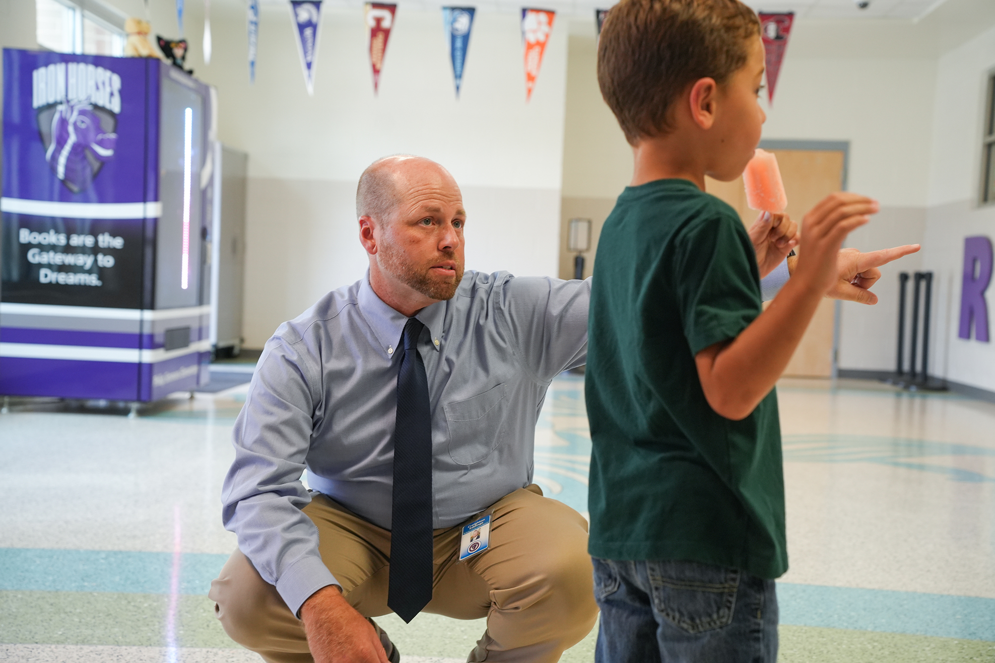 A man kneels with a child in a school hallway, both holding popsicles. Behind them, a refrigerator is visible.