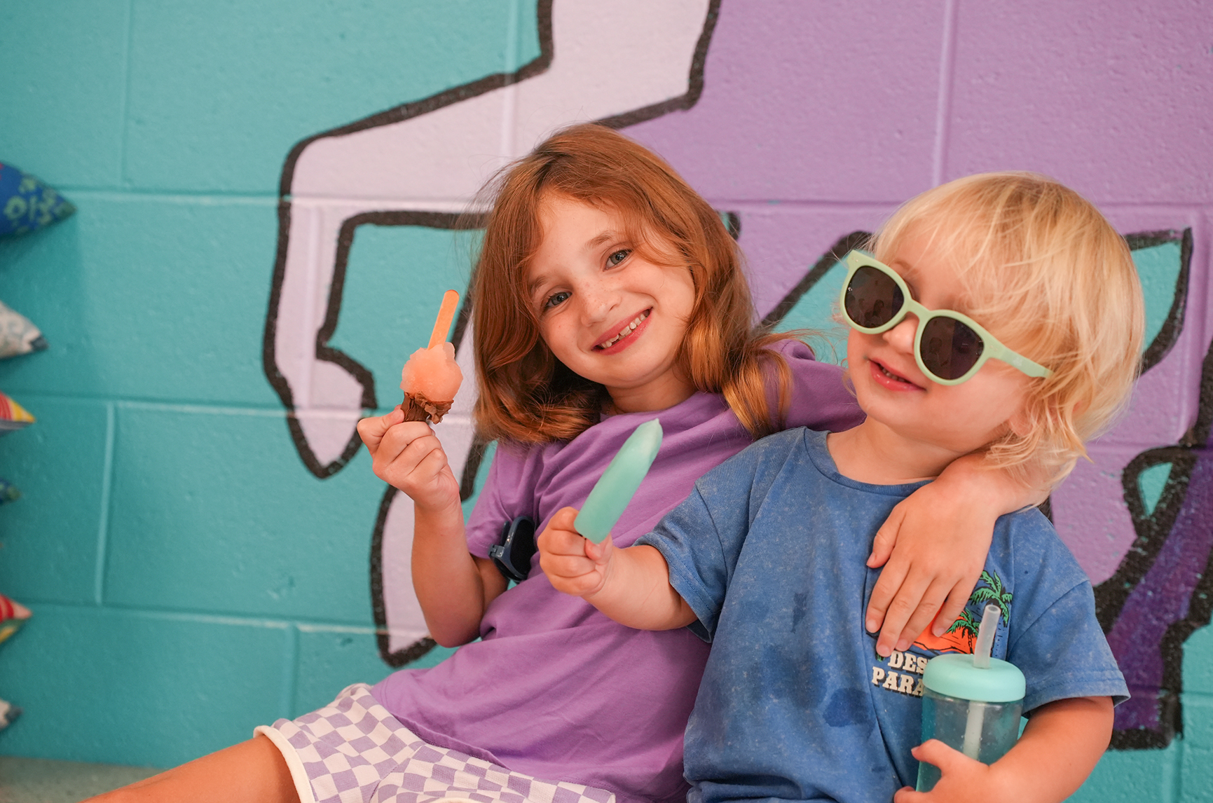 Two children, one with a pink shirt and the other in blue, smile while holding ice pops. They sit in front of a colorful mural.