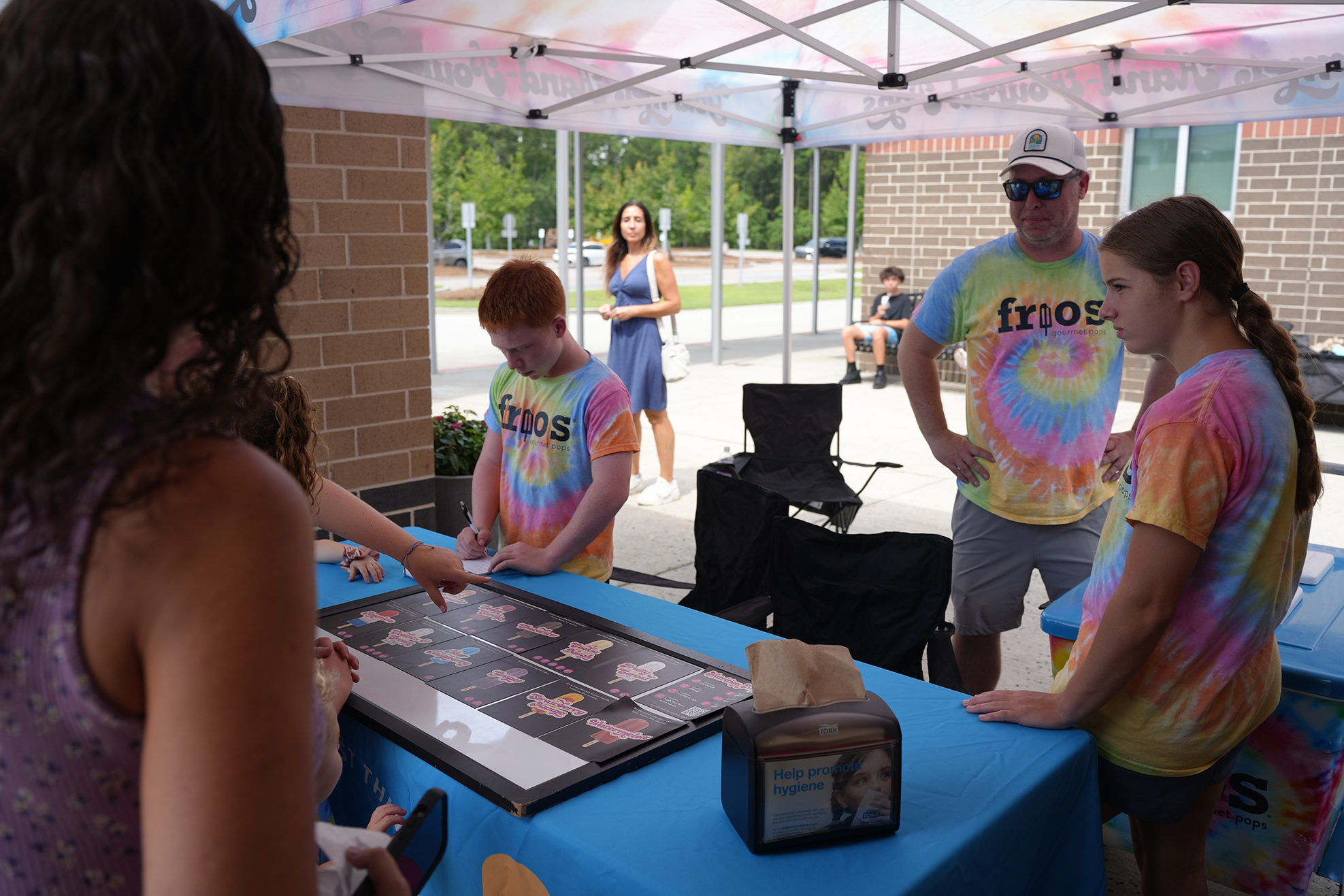 People in tie-dyed shirts at a table under a tent. One person points at a framed photo.