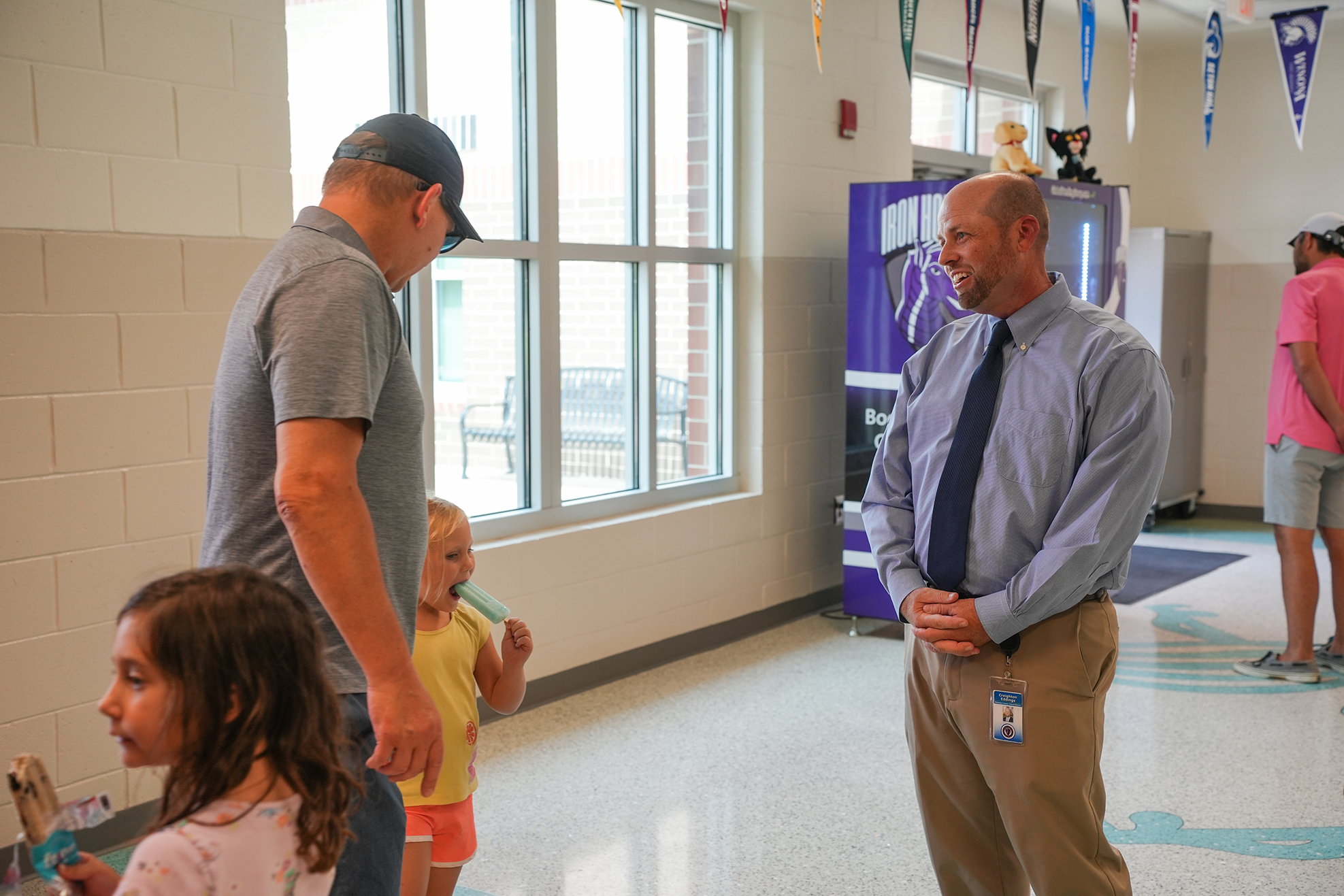 Two men and two children stand in a hallway, one man wearing a cap and the other in a tie. A vending machine is nearby.