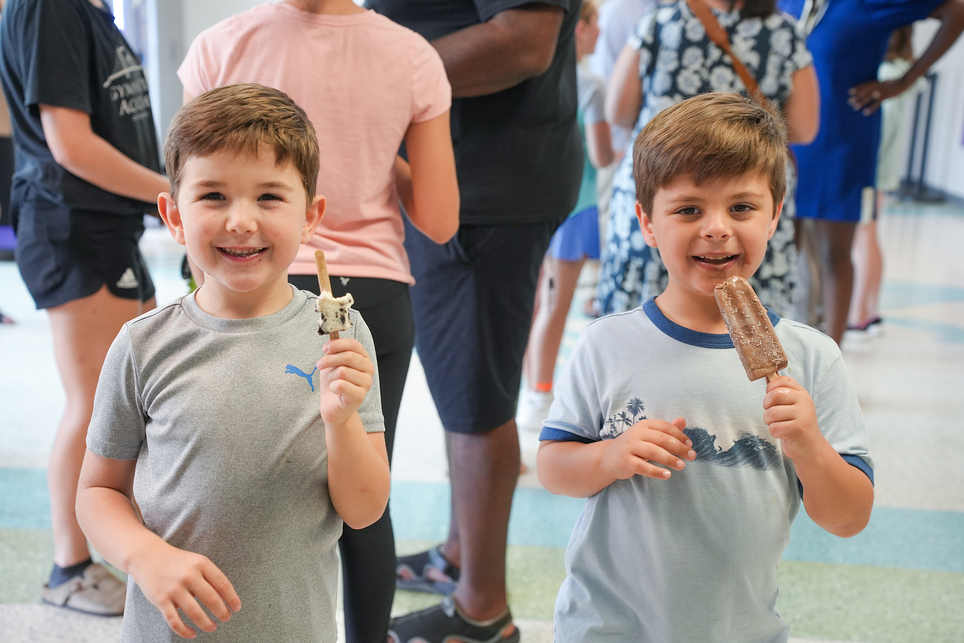 Two boys, both smiling, each holding a popsicle in their right hand, stand in an indoor area with others.