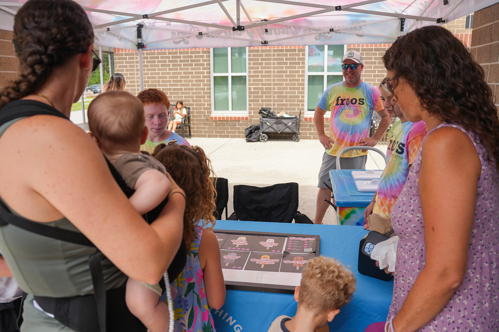 Several adults and children gathered around a table, one woman holding a baby. Background includes a building.