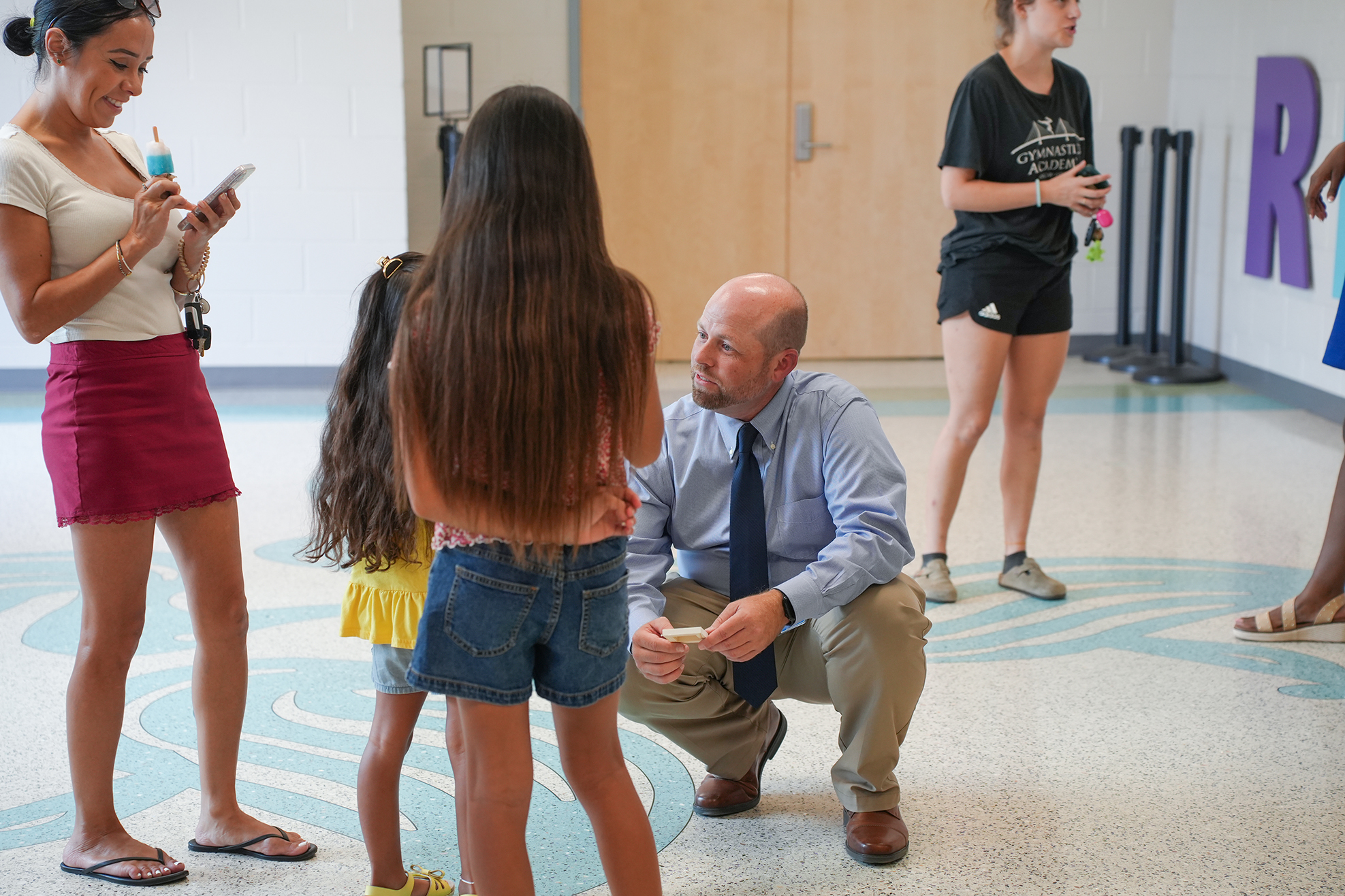 A man kneels, speaking to a girl. Women in shorts and dresses surround him. A purple letter R is on the wall.