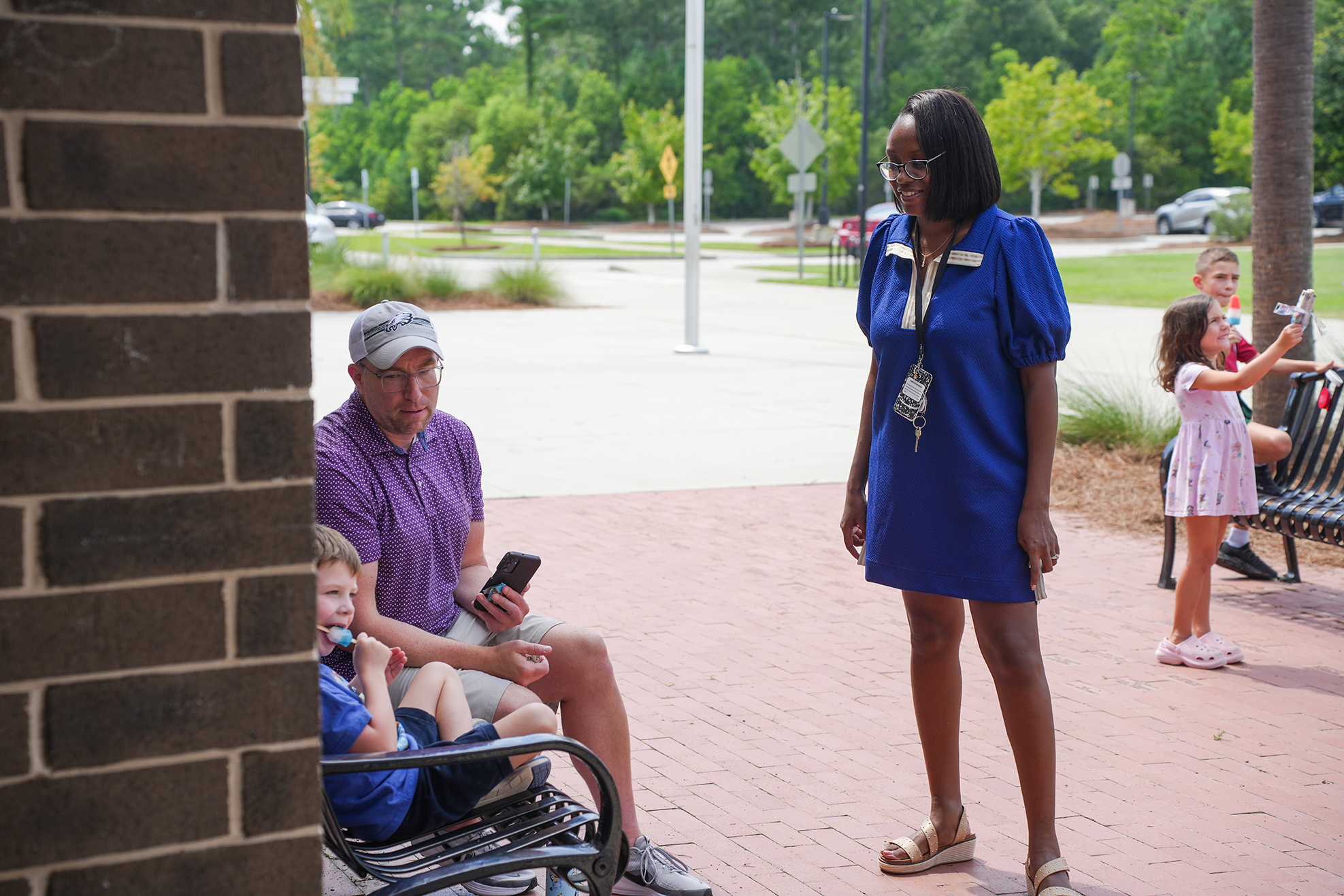 A woman stands by a bench; a man sits on the bench with two children, one looking at a phone.