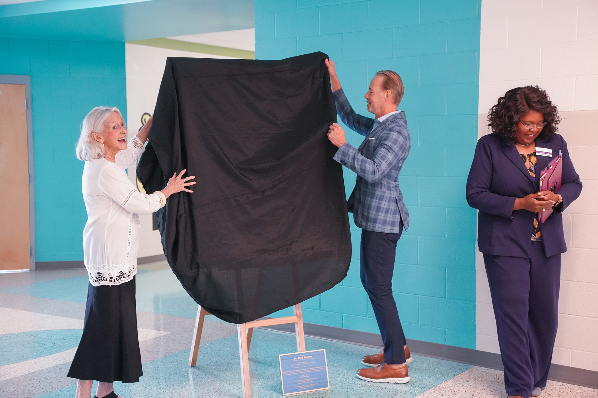 Three people unveil a large black cloth in a hallway. The woman on the left wears a white top.