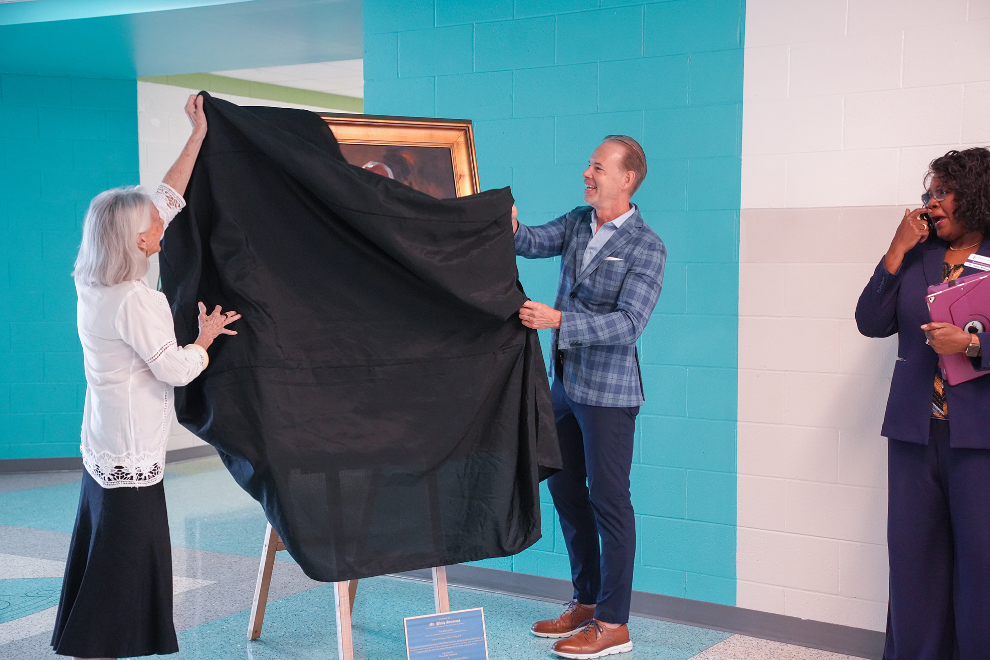Two people unveil a large black cloth from a framed picture. A third person stands nearby. A white wall and blue-painted wall are in the background.