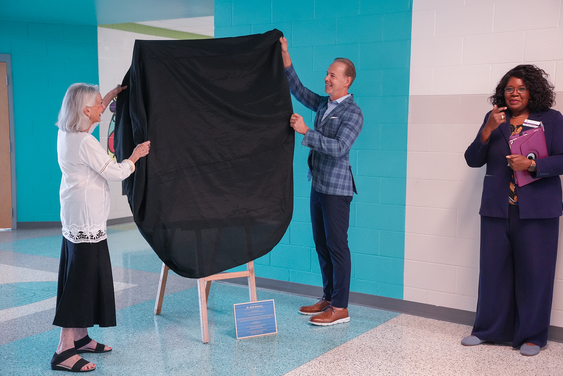 Three people hold a covered item in a hallway. A woman on the left holds the fabric up. A man and another woman hold the sides.
