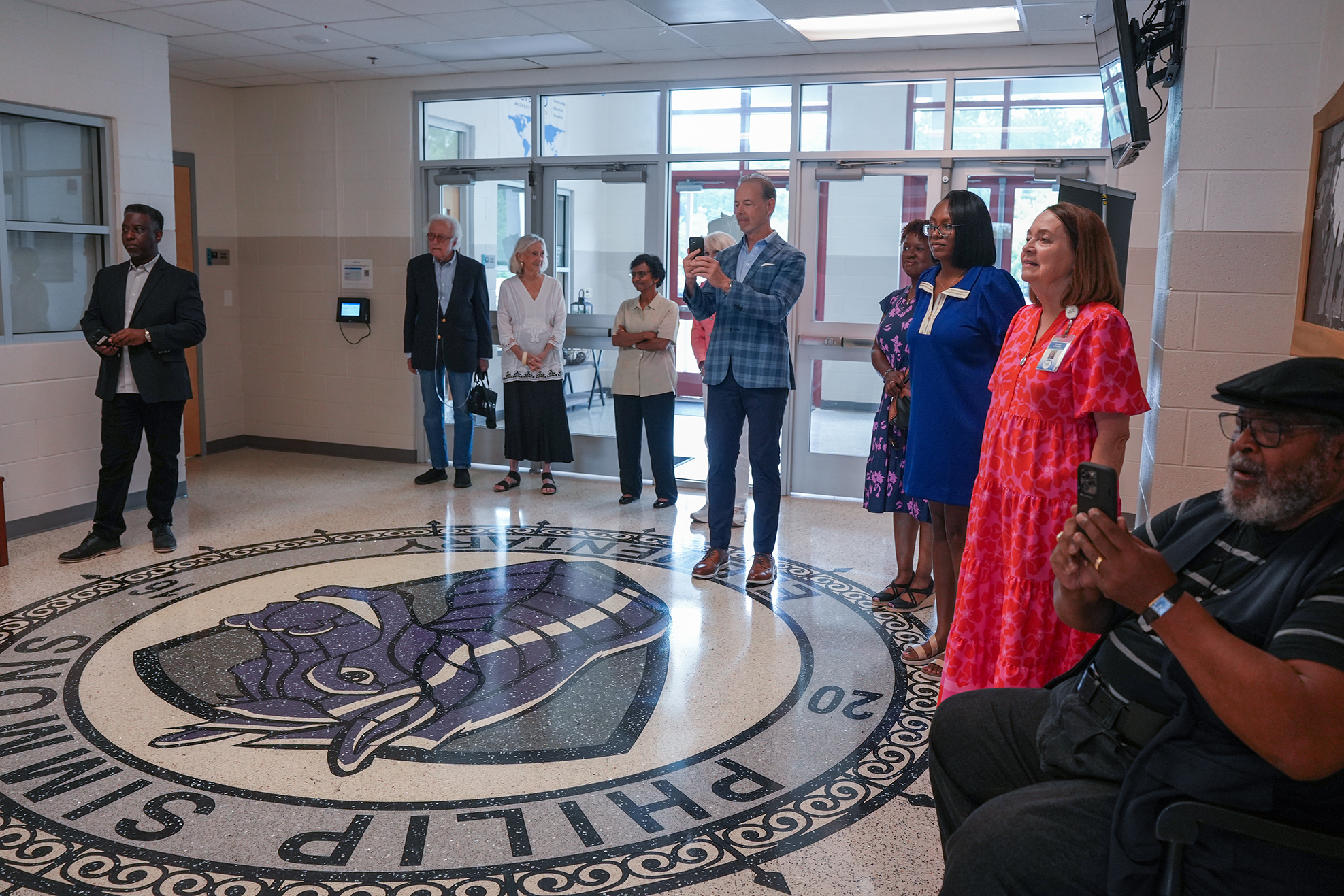 A group of people stand around a school logo on the floor. A person takes a photo.