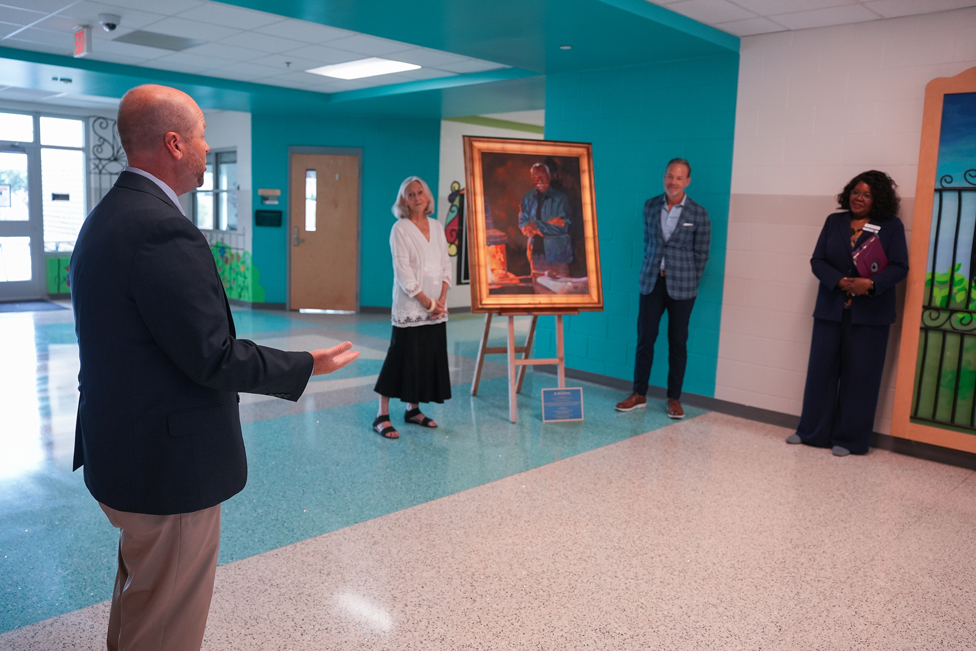 Four people stand in a room with blue walls, a painting on an easel, and a window.