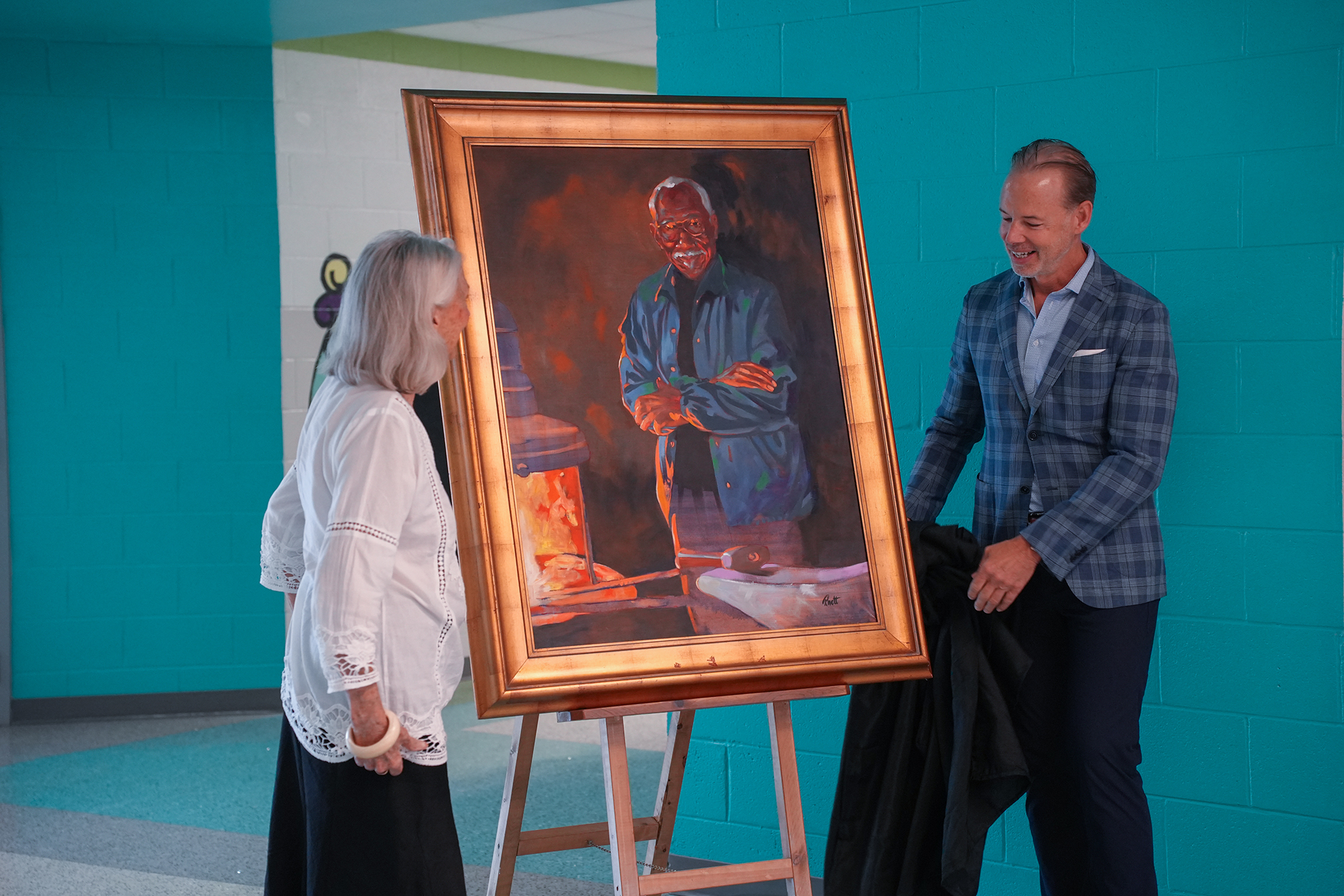 A woman in a white top and a man in a blue suit stand near a portrait of a man.