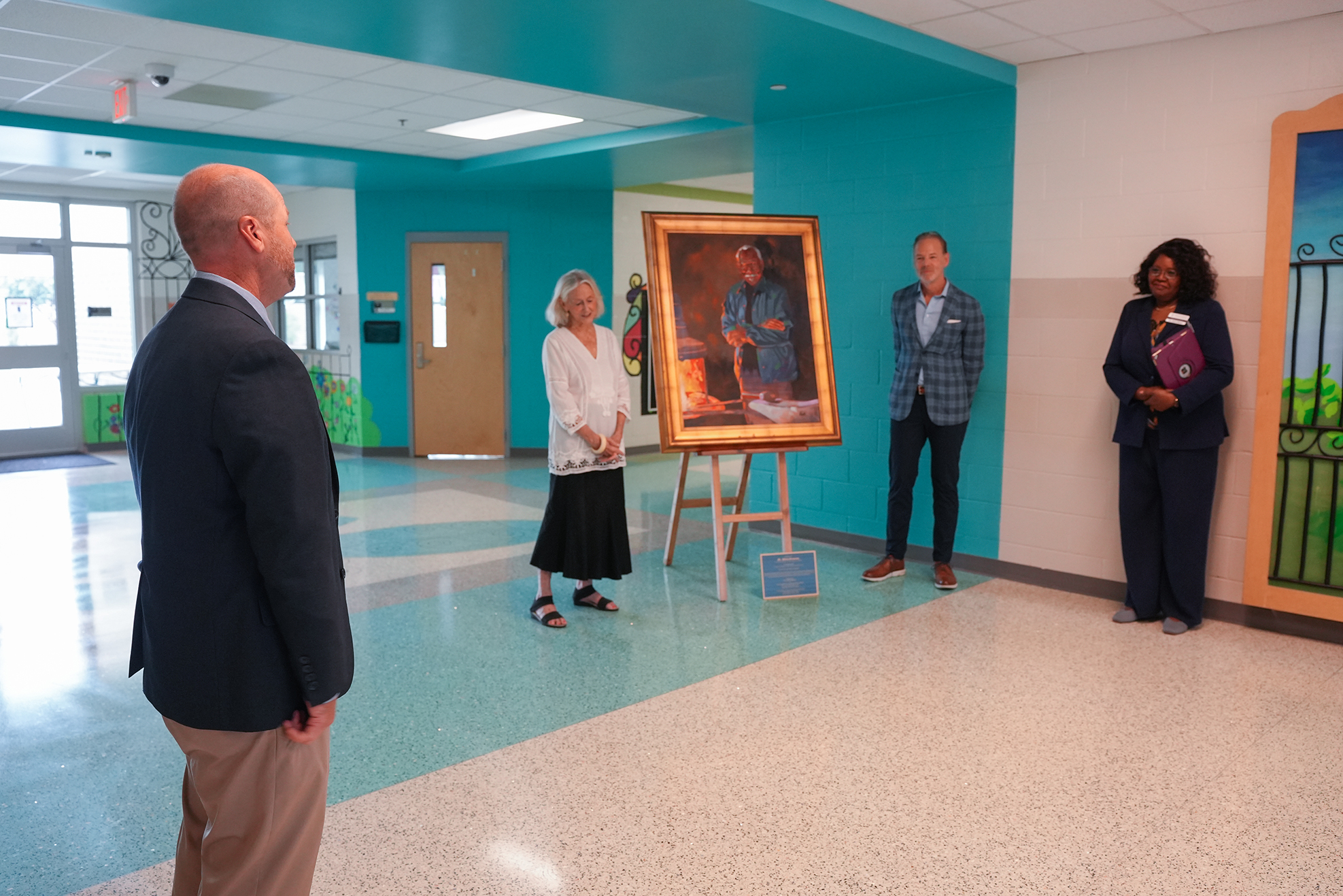 A group of people stand in a hallway. One woman holds a framed portrait on an easel.