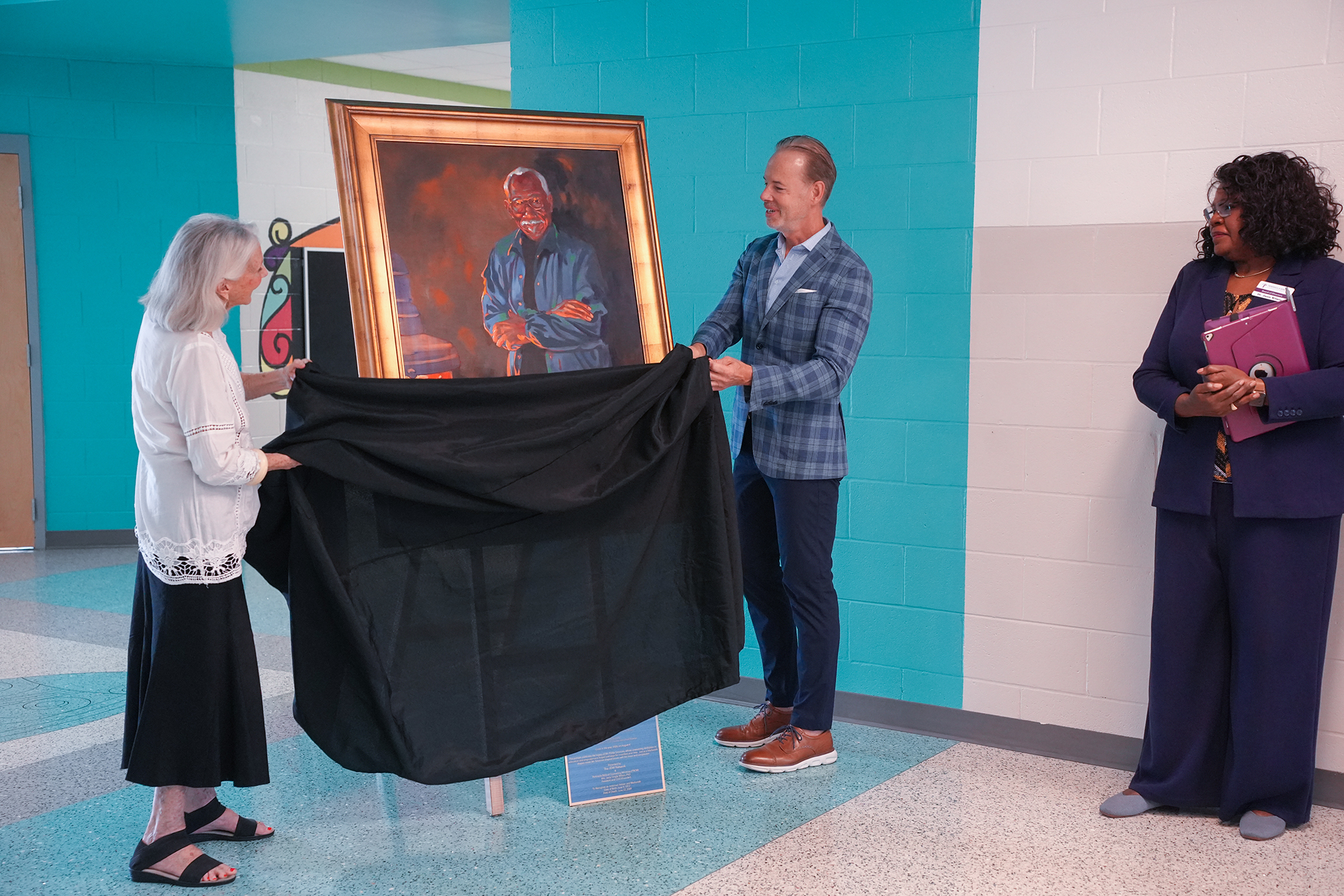 Three people unveil a portrait on a stand in a hallway. Two men and one woman are present.