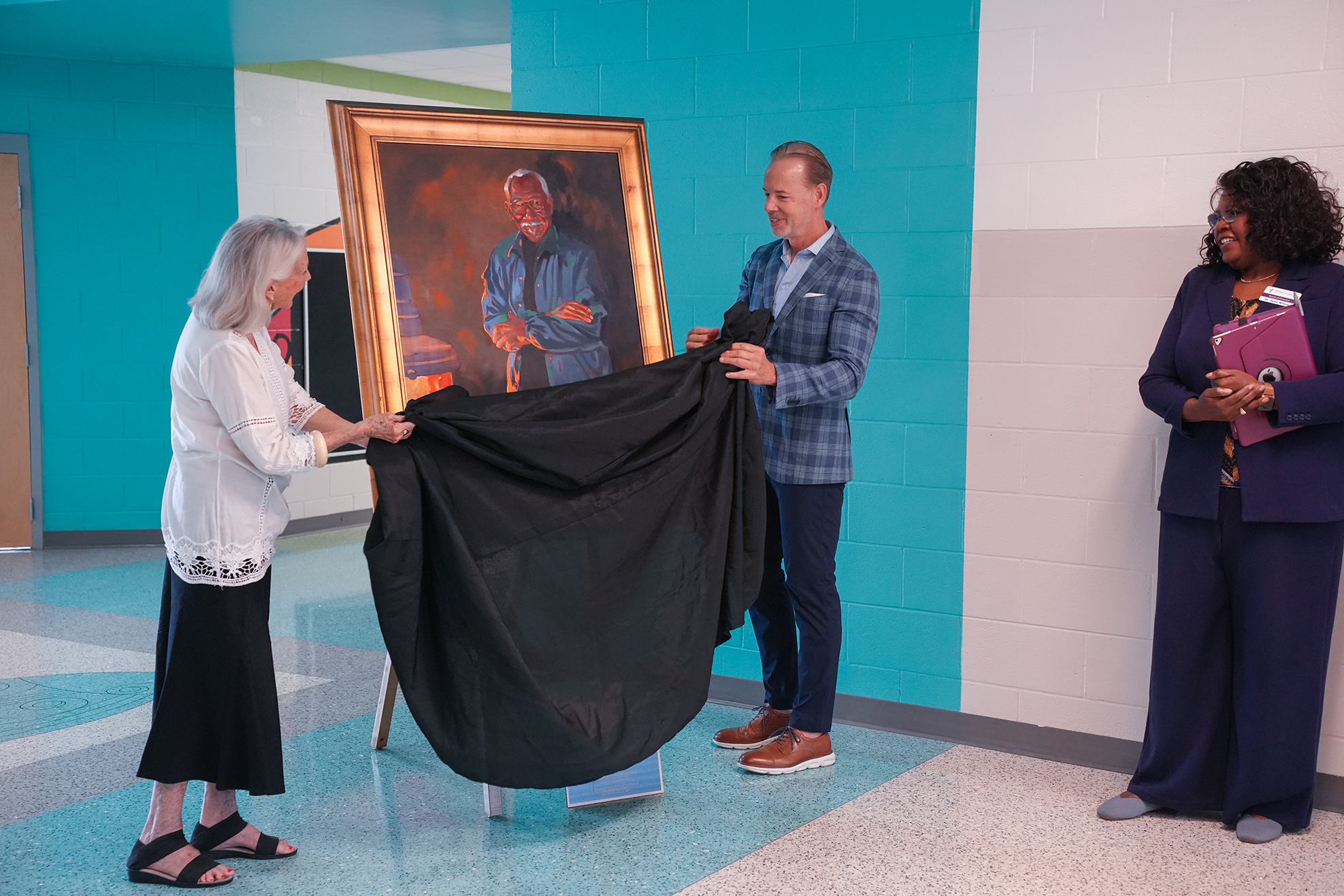 Three individuals unveil a portrait in a hallway. The man on the right holds a folder, the woman on the left holds the cloth, and a man in the middle lifts the frame.