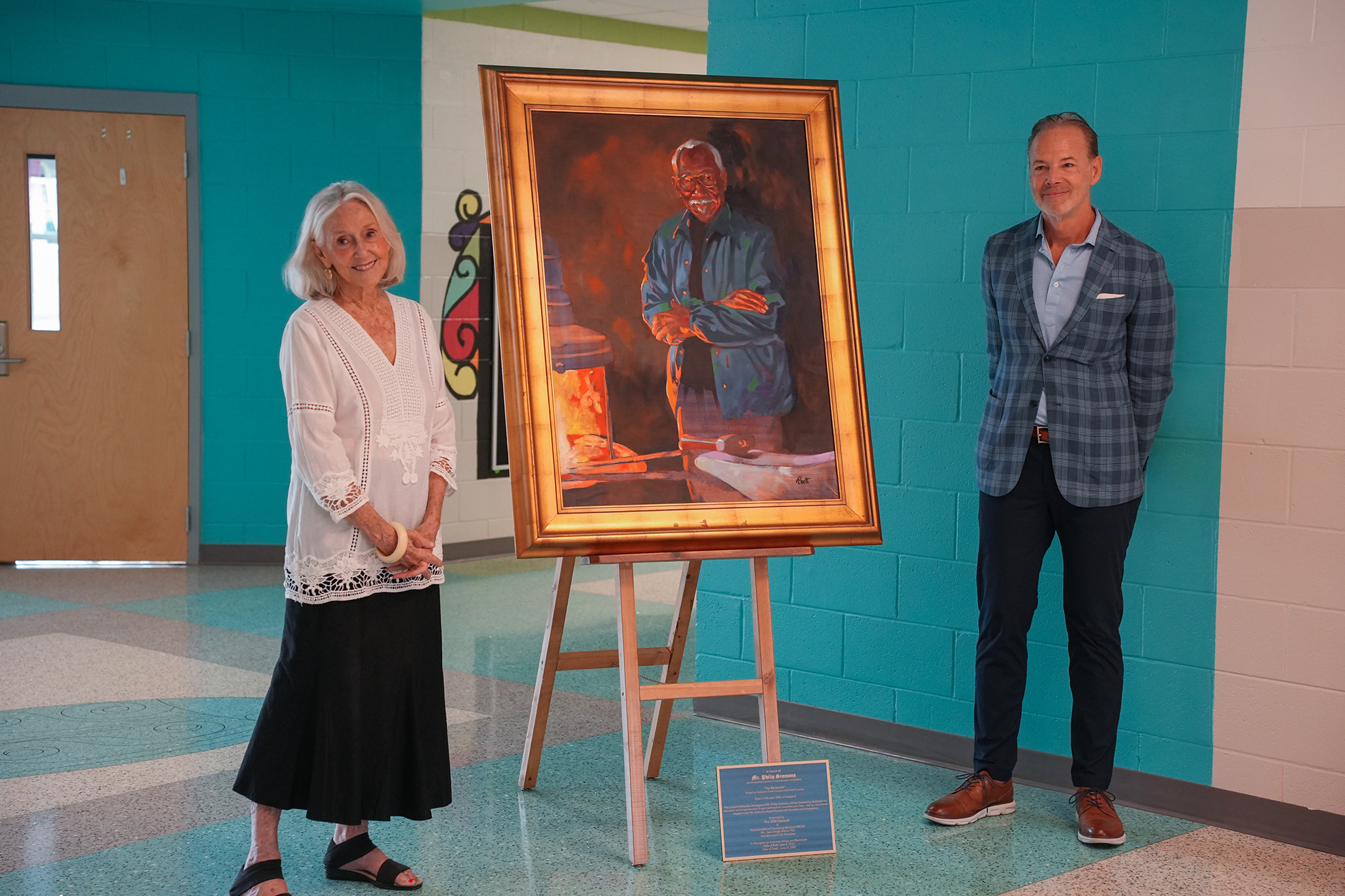 Two adults stand next to a portrait painting on an easel in a hallway. Blue walls and a wooden door are visible.