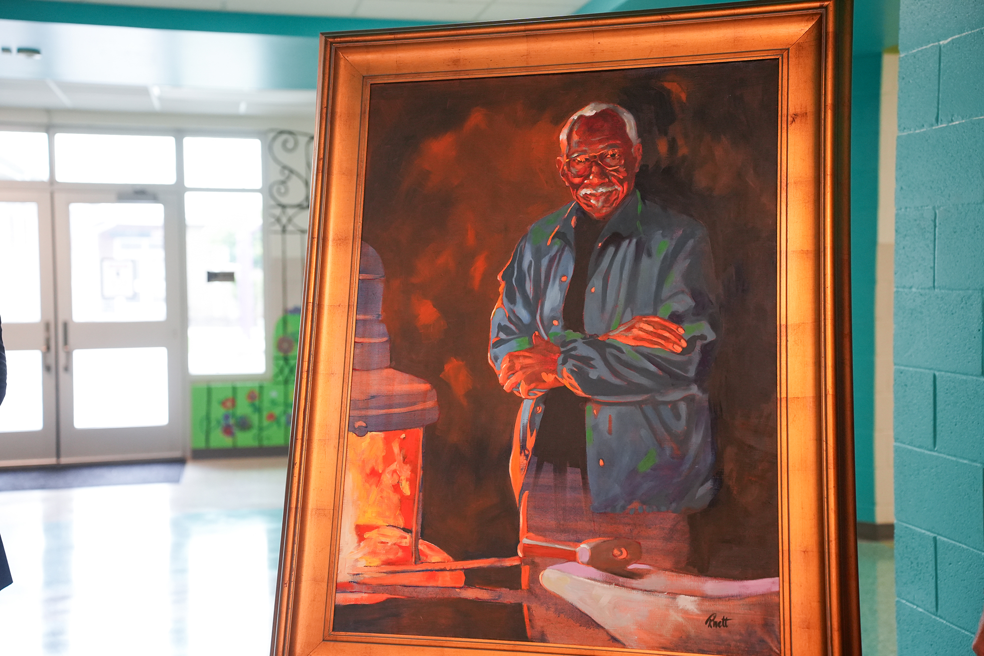 A framed painting depicts a man standing with arms folded. The painting has warm colors and shows a workshop background.