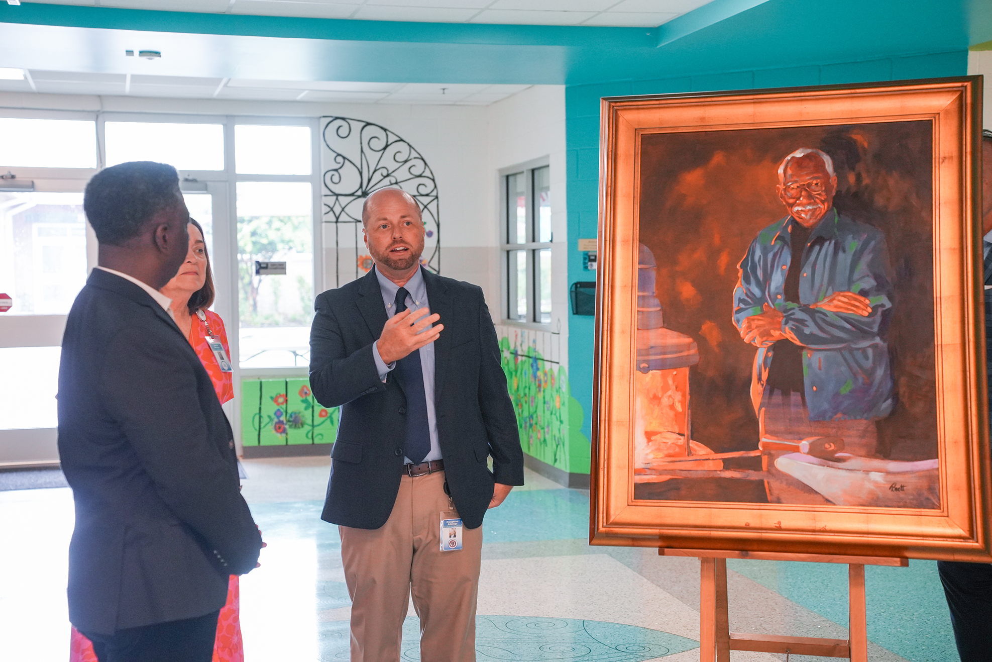 A man in a suit speaks next to another man at an exhibit. The painting is of a man in a blue jacket.