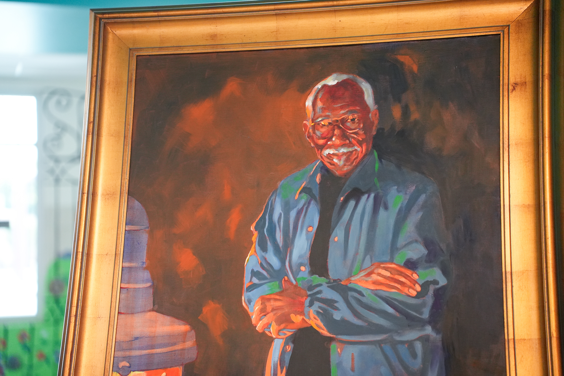 Portrait of a man with white hair and glasses, wearing a blue shirt, standing with folded arms. Framed in a gold frame. Background includes a window.