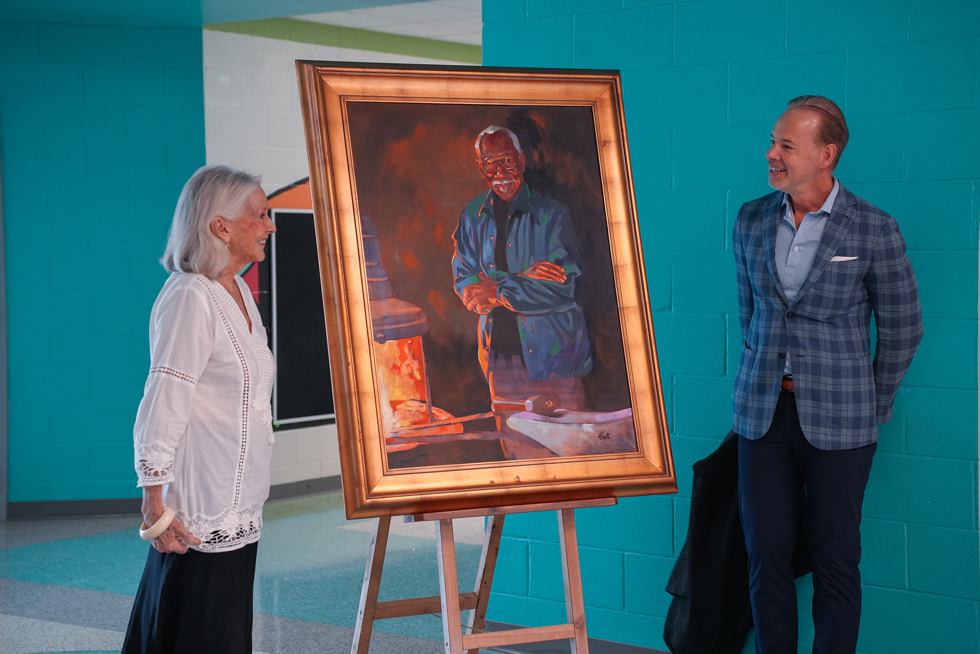 Two people stand before a framed painting, one person in white, the other in a plaid jacket.