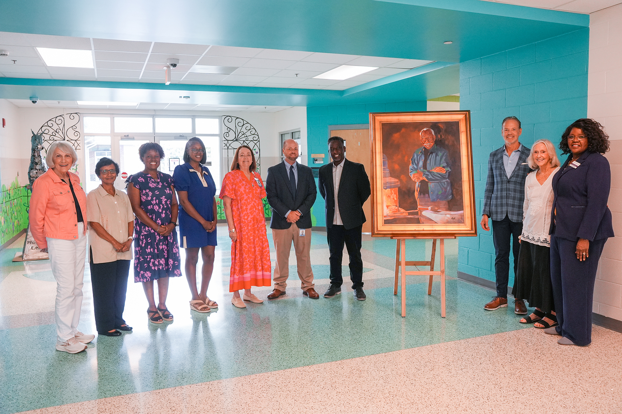 A group of adults in formal and casual clothing stands around a portrait on an easel in a brightly lit hallway.